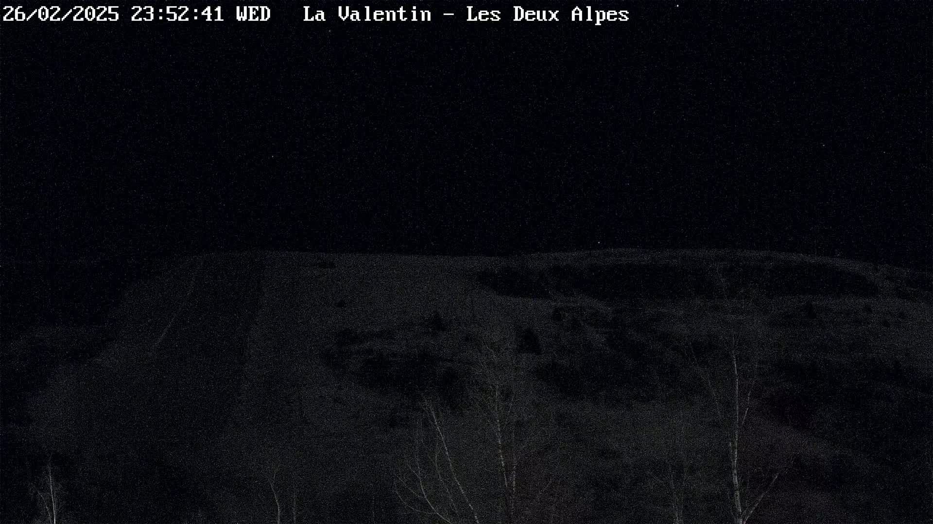 A dark, nighttime view of a snow-covered hill with sparse, dark trees in the foreground.
