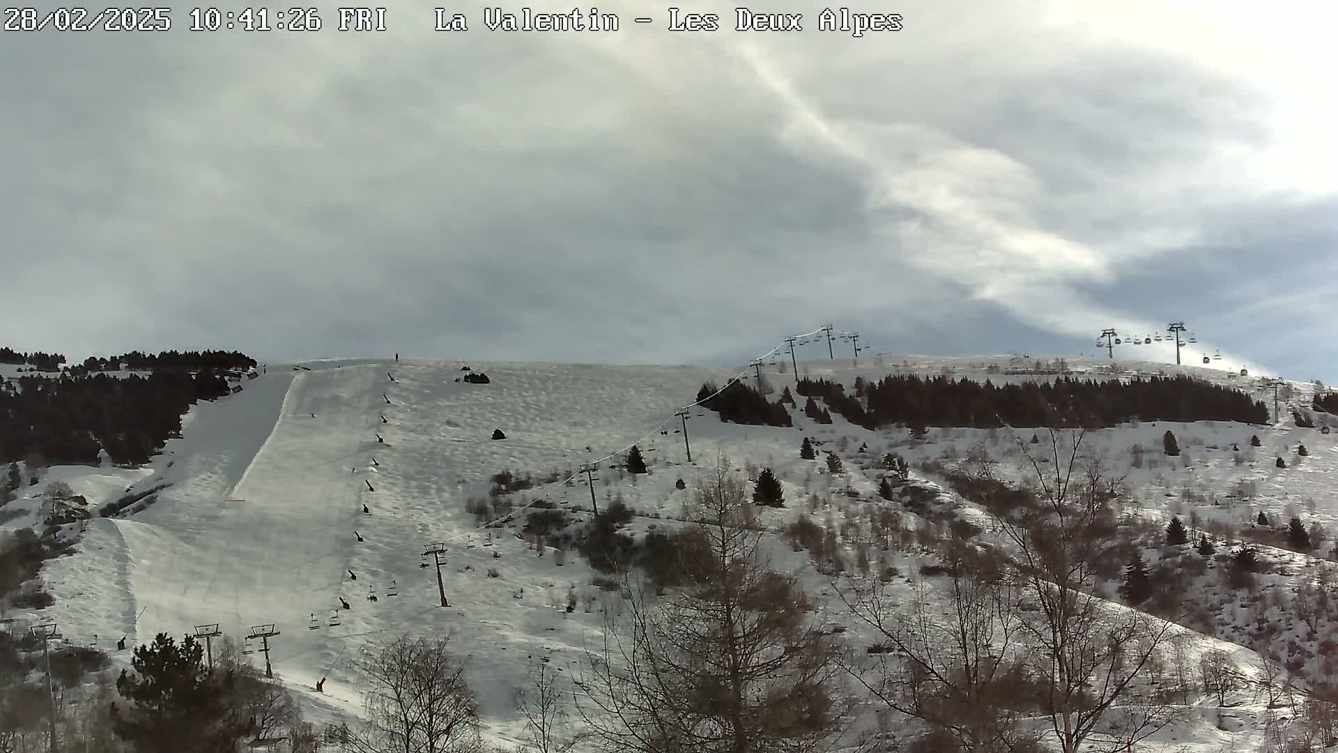 A snow-covered ski slope with chairlifts and sparsely wooded areas under a mostly cloudy sky.