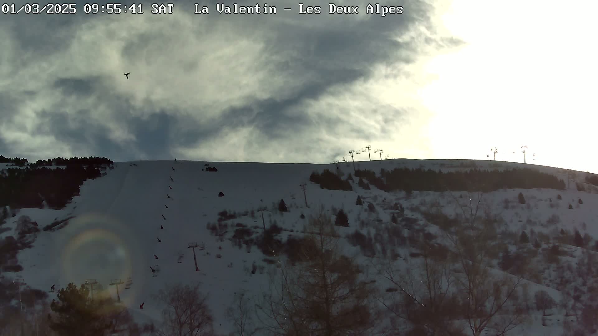 A snow-covered hillside with ski lifts and sparsely populated trees under a partly cloudy sky.