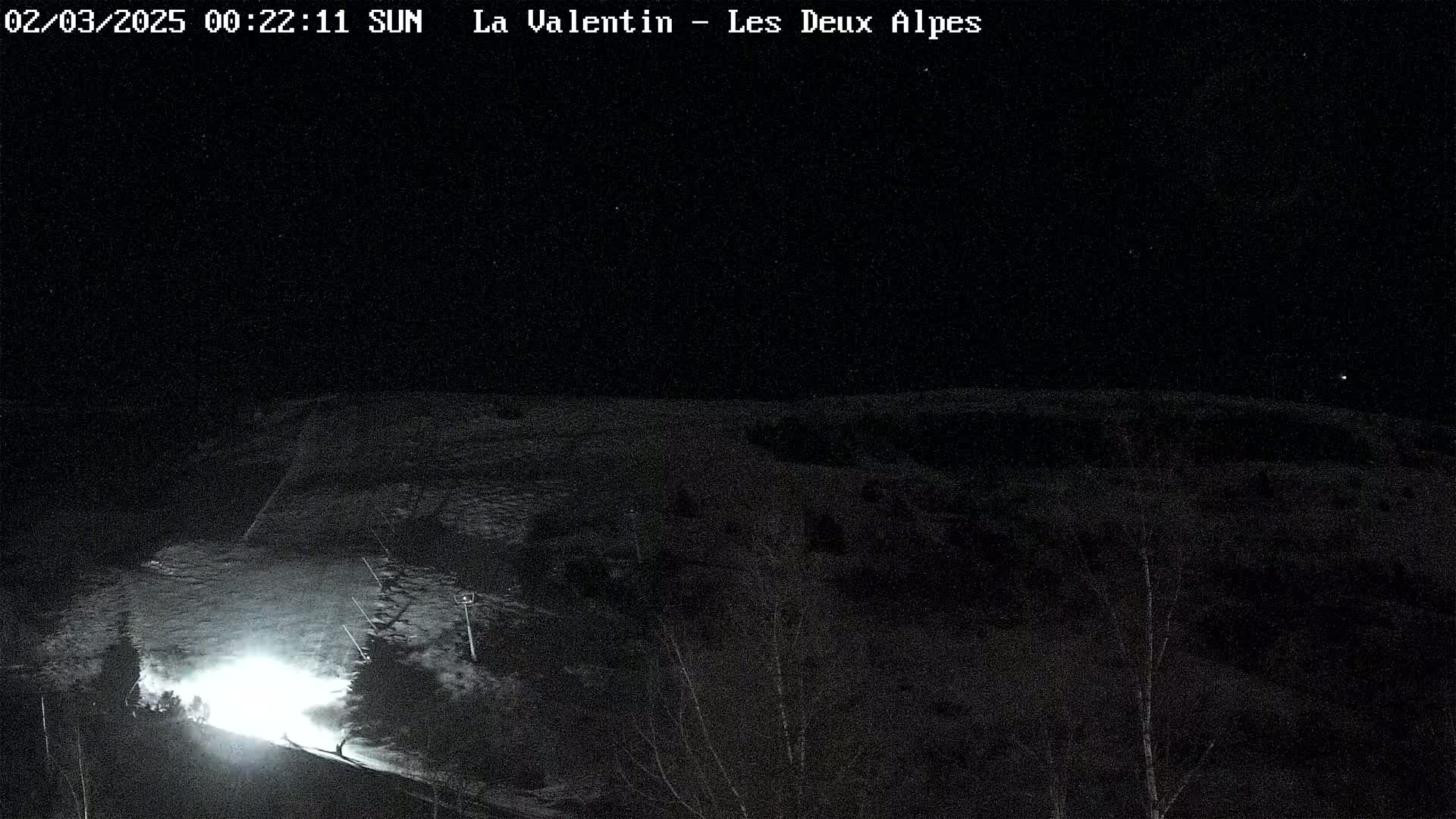 A nighttime, snow-covered landscape is illuminated by a bright light source at the bottom, with dark trees and hills visible in the background under a clear, starlit sky.
