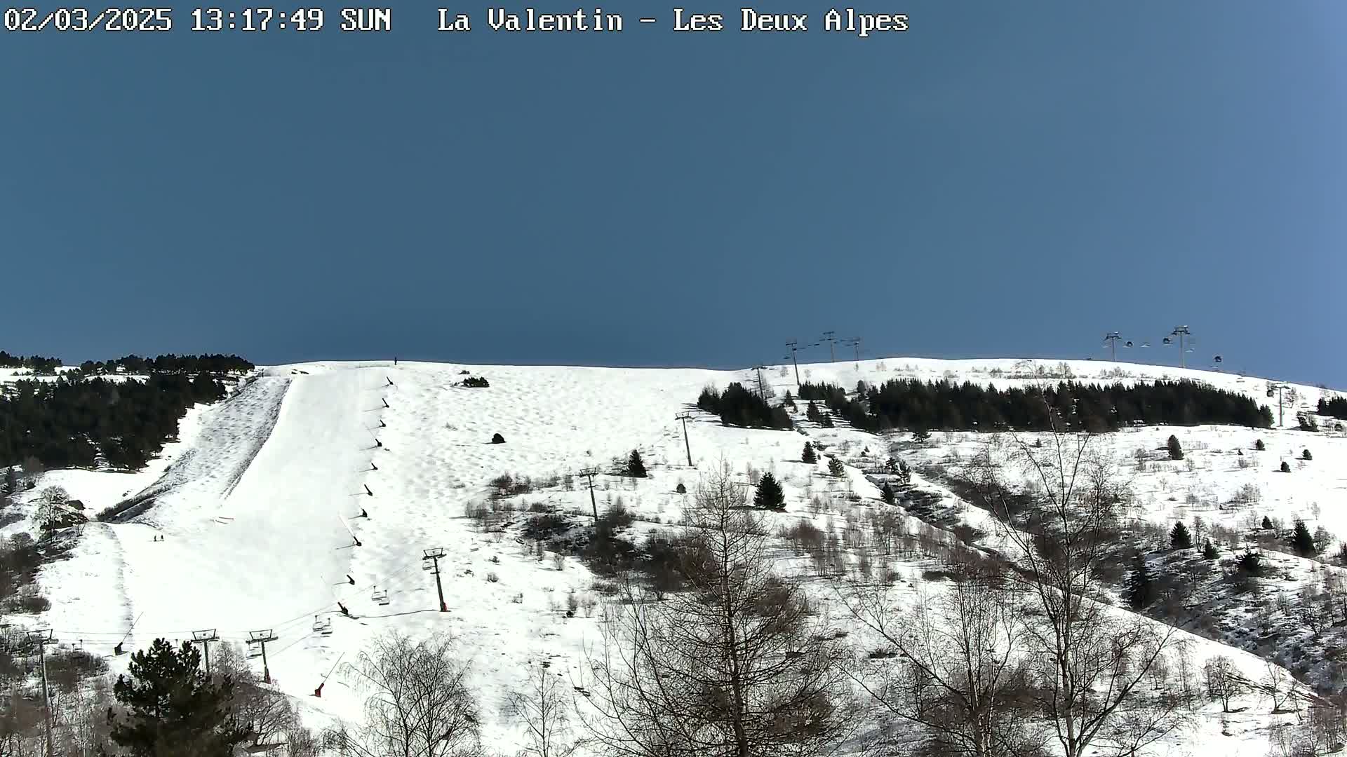 A snow-covered ski slope with chairlifts and sparsely-treed hills under a clear blue sky.