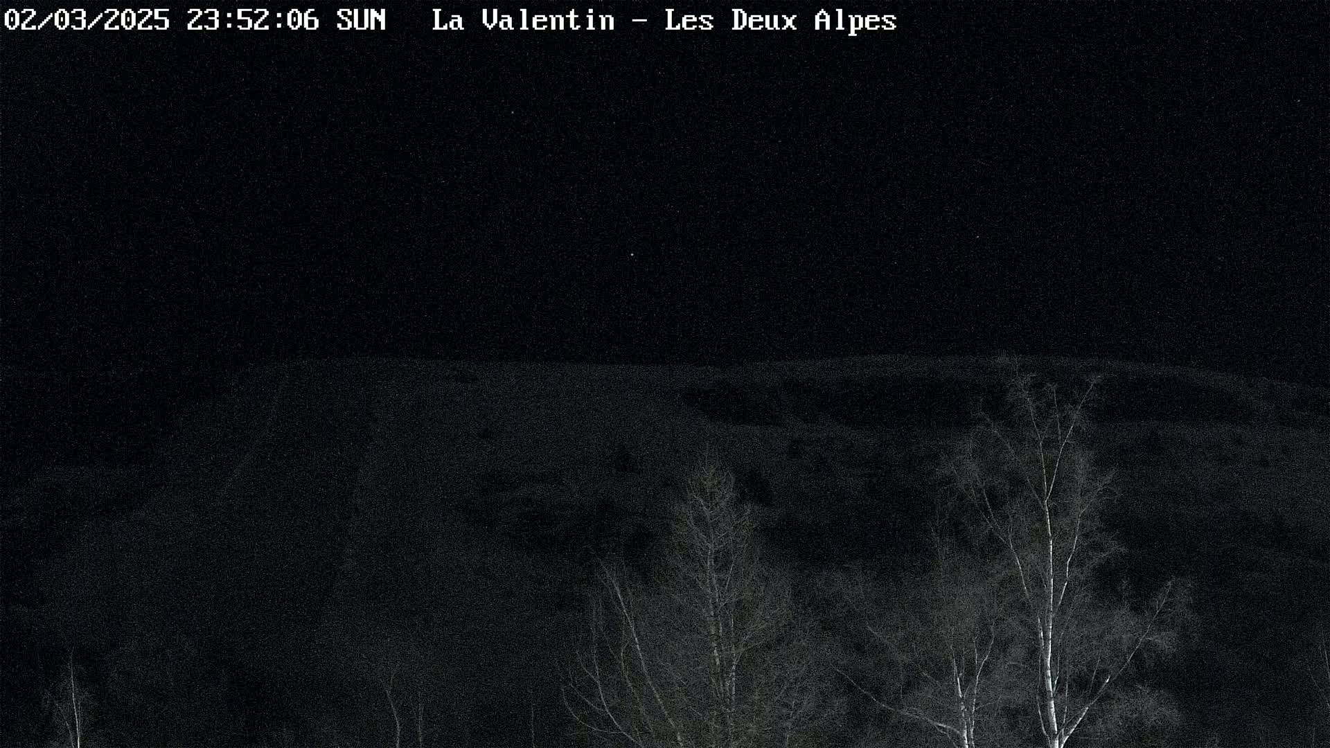 A nighttime view of a snow-covered landscape with leafless trees in the foreground under a clear, dark sky.