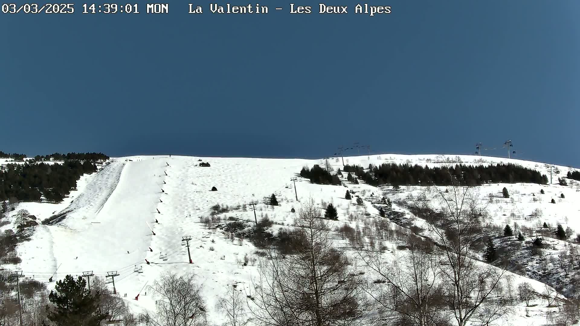 A snow-covered ski slope with sparsely wooded areas and ski lifts under a clear blue sky.