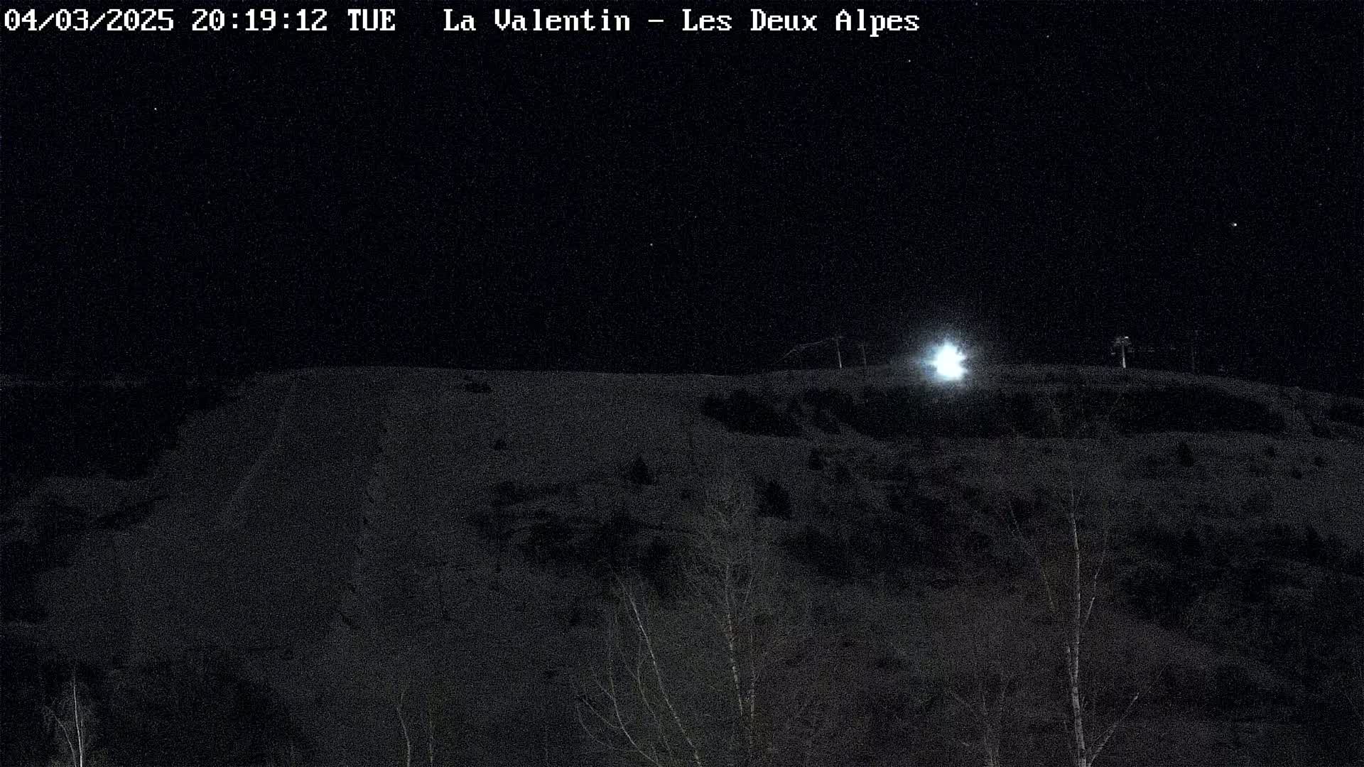 A nighttime, snow-covered hillside is illuminated by a bright light source, with sparse, dark trees scattered across the landscape.