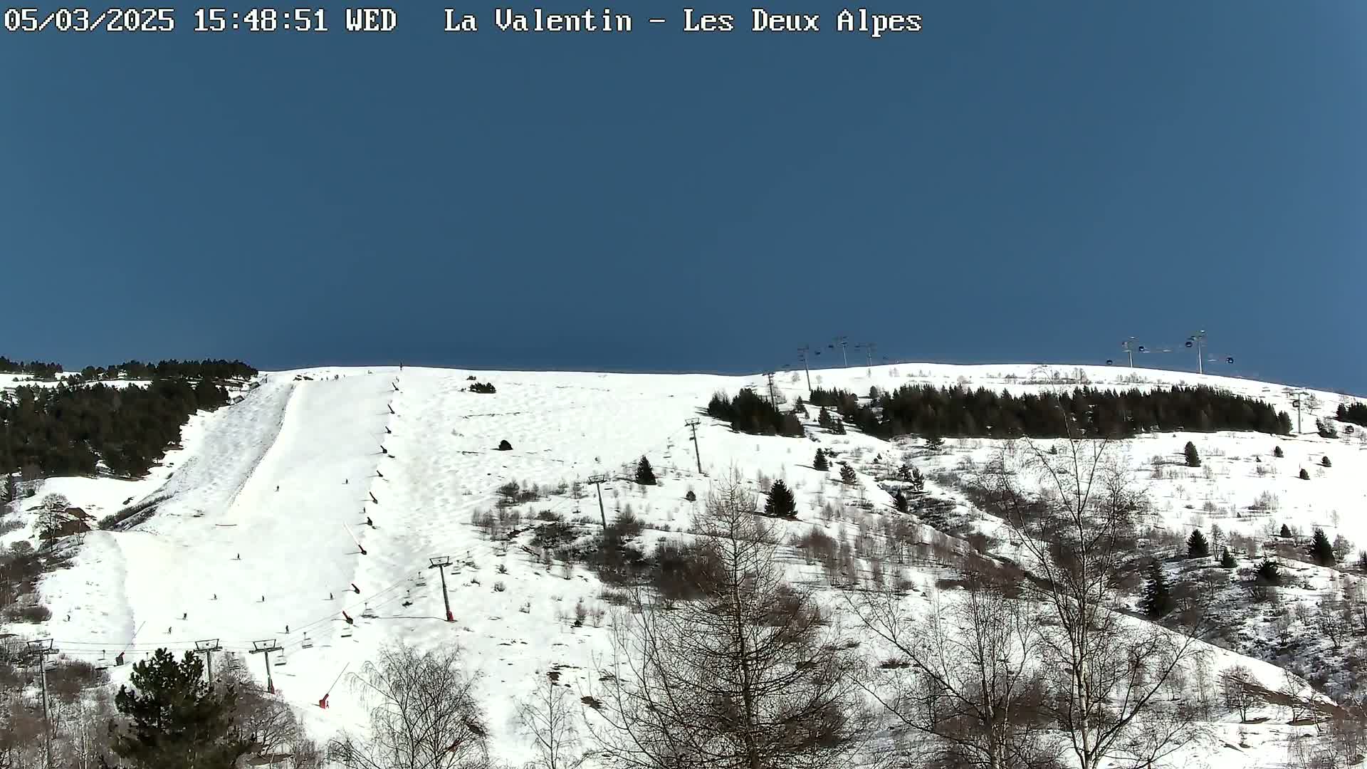 A snow-covered ski slope with sparsely treed hillsides under a clear blue sky shows several skiers and ski lifts.