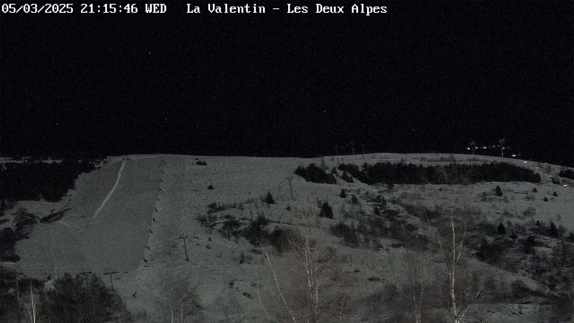 A snow-covered hillside at night, with sparsely wooded areas and a ski slope illuminated by lights.