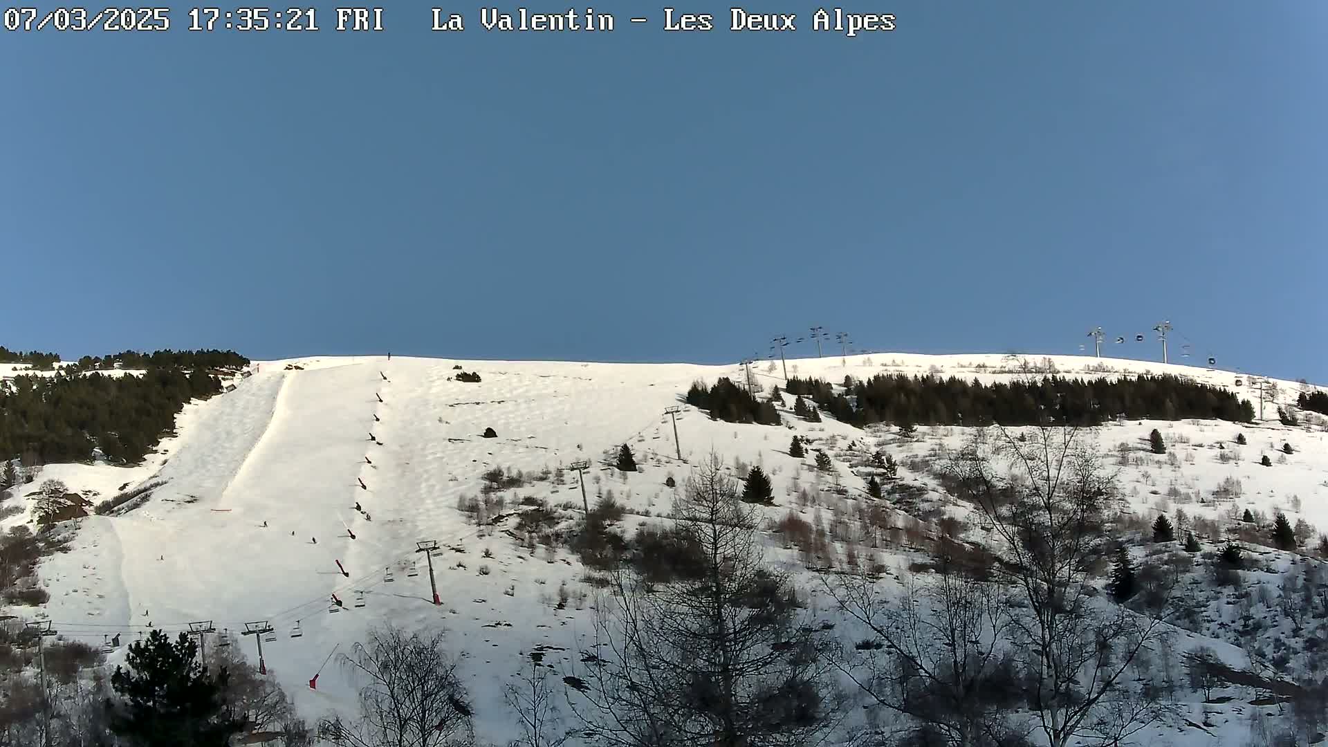 A snow-covered ski slope with sparsely wooded areas and ski lifts under a clear blue sky.