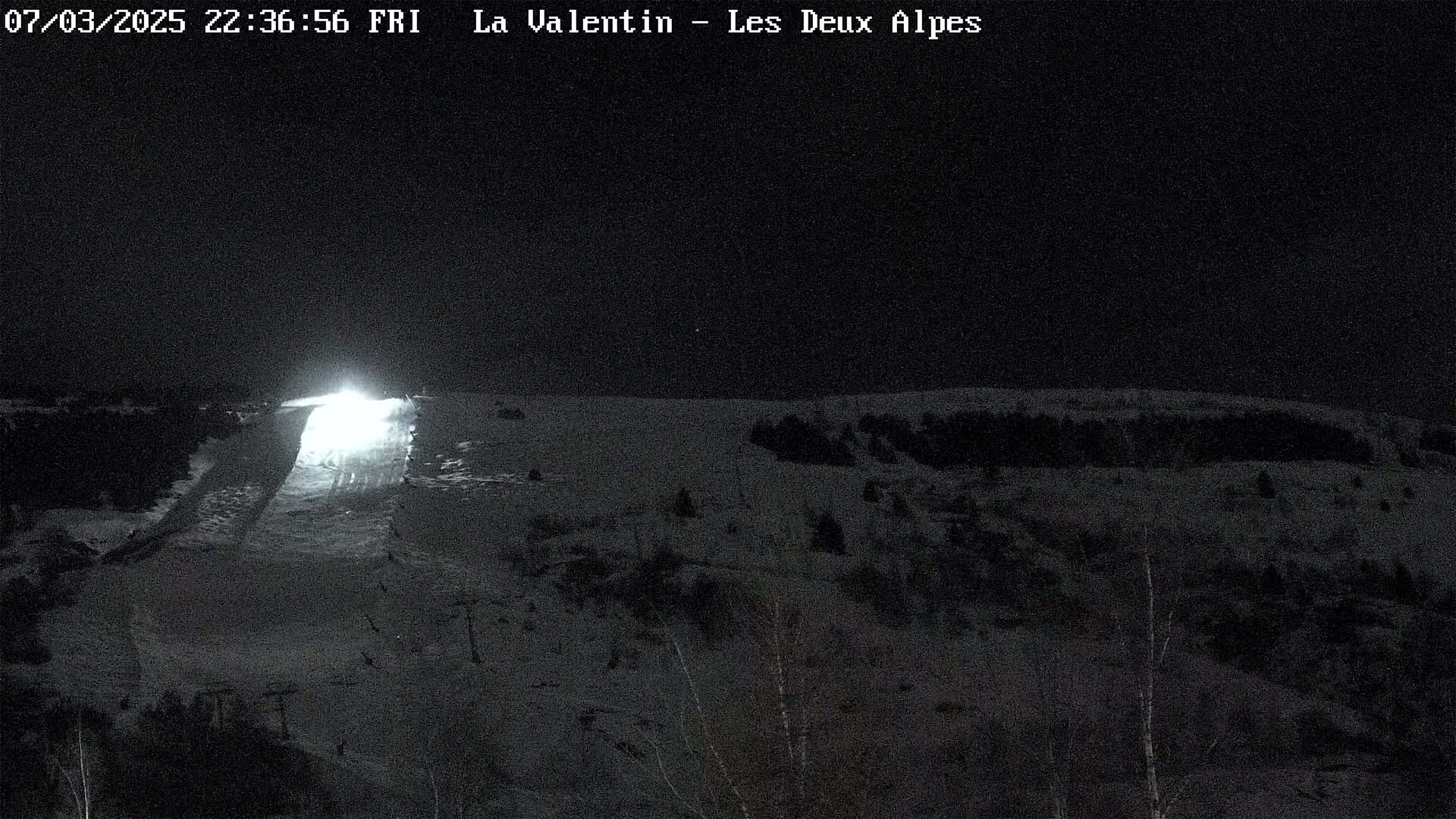 A brightly lit snow-covered slope is visible at night, with dark trees and a snowy landscape surrounding it.