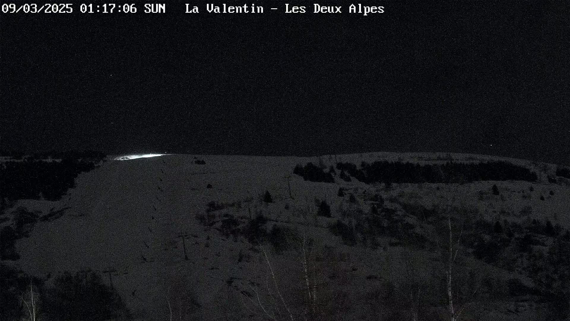 A nighttime, snow-covered landscape with sparsely scattered trees and faint lighting in the distance.