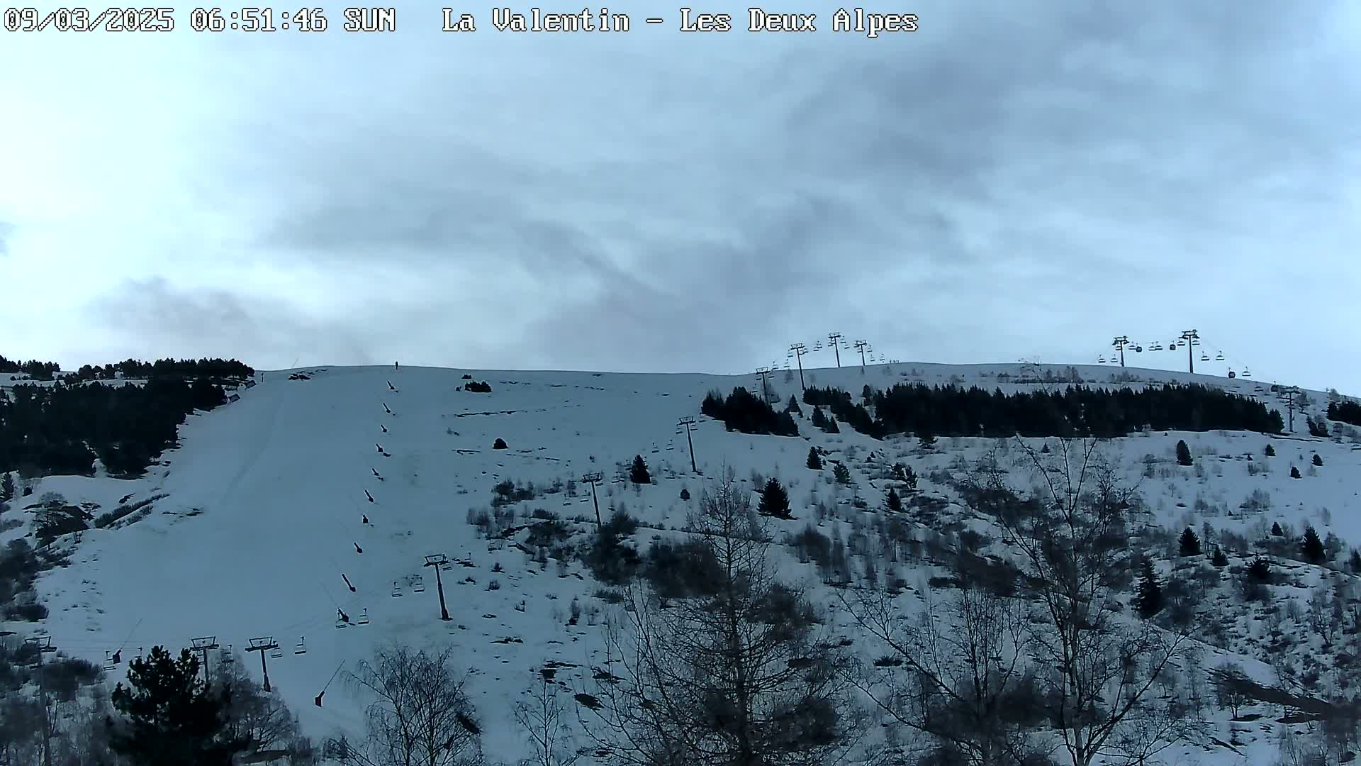 A snow-covered ski slope with sparsely treed hillsides and chairlifts under a cloudy sky.