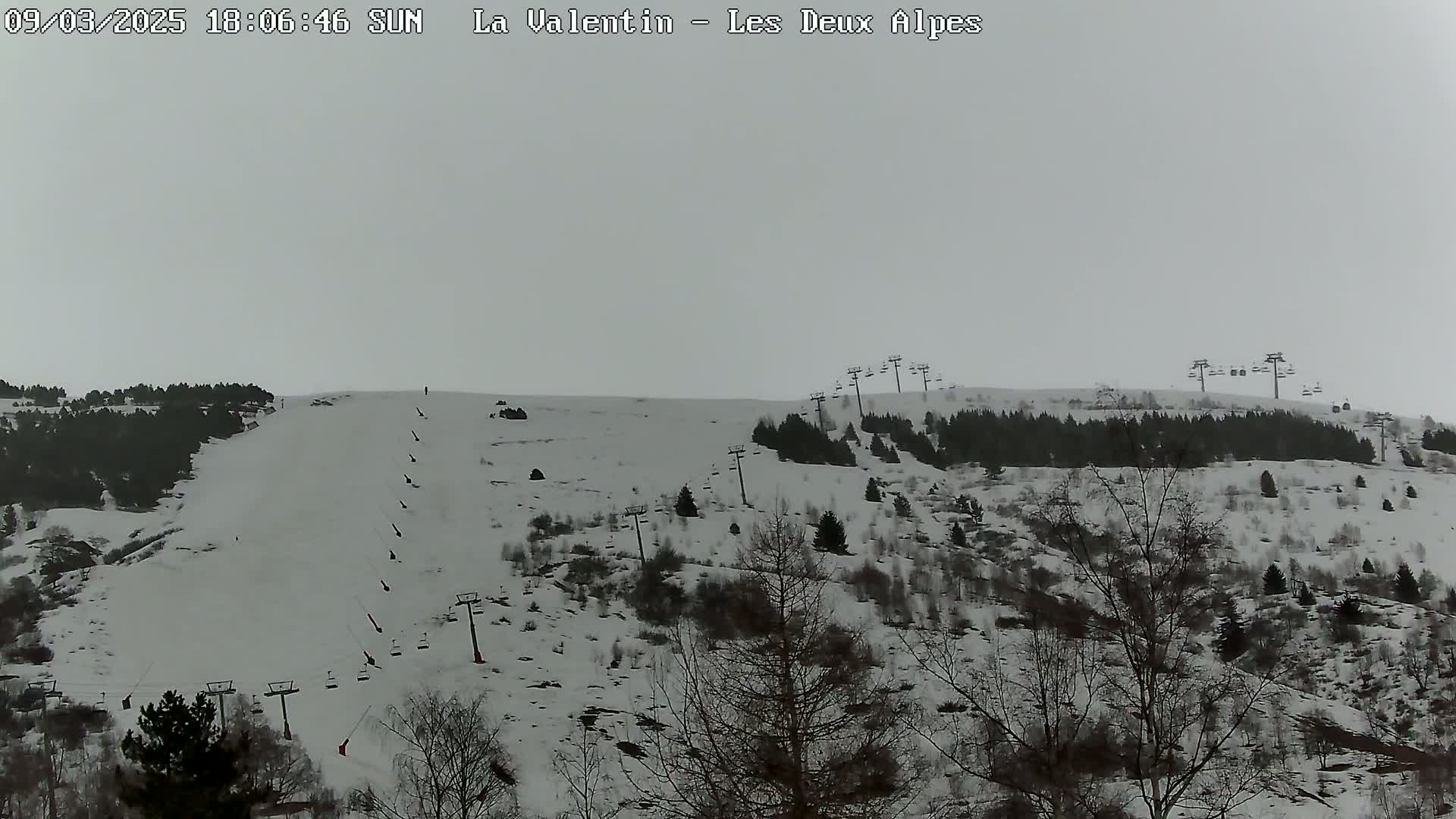 A snow-covered hillside with sparsely wooded areas and several ski lifts under an overcast sky.