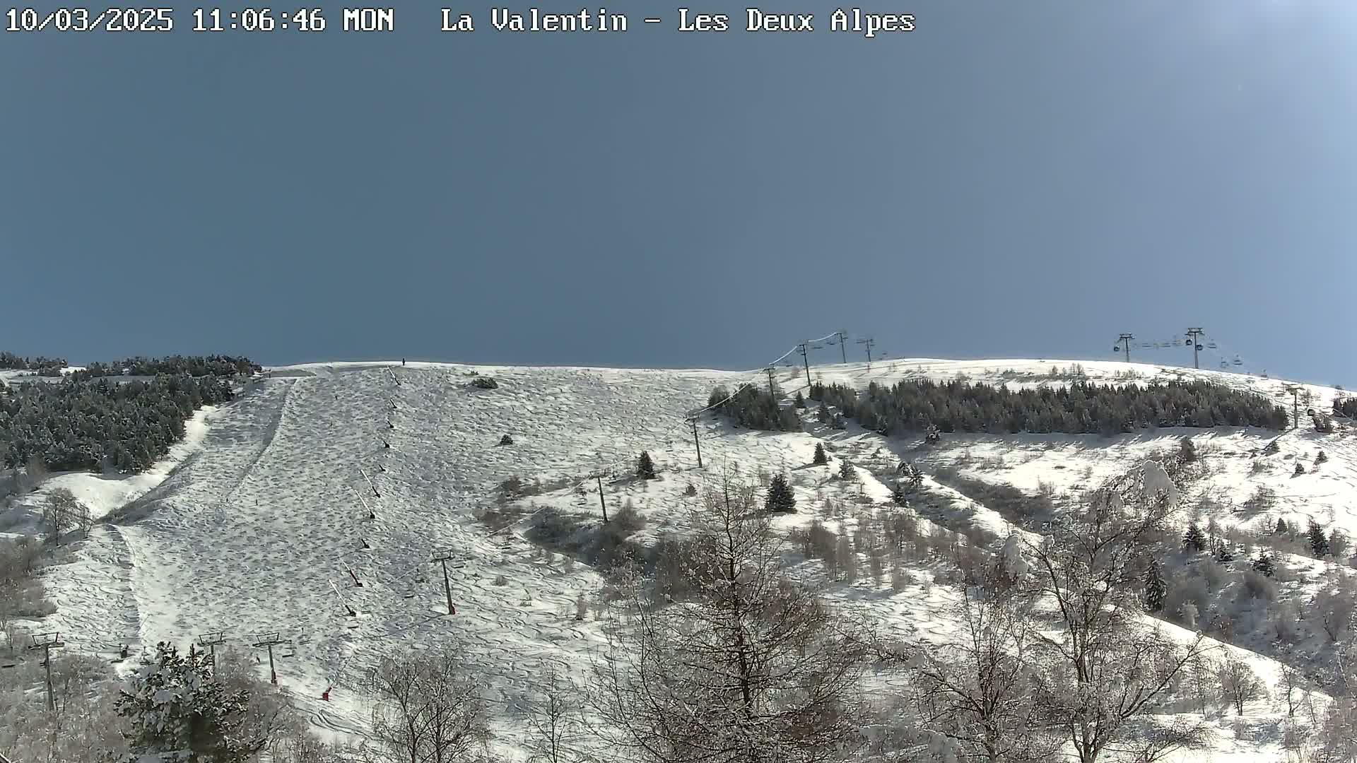 A snow-covered mountain slope with ski lifts and sparsely wooded areas under a clear blue sky.