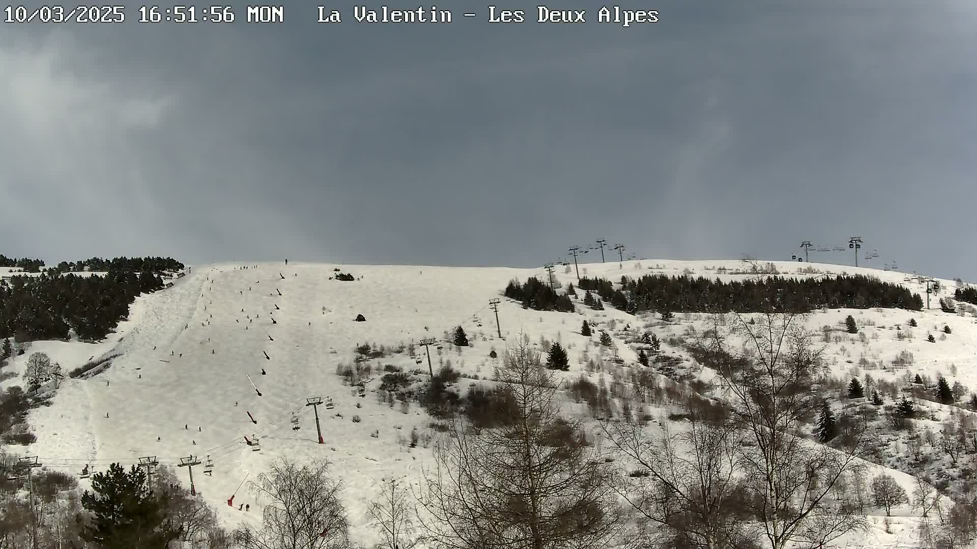 A snow-covered ski slope with several skiers visible, under a cloudy sky, is surrounded by snow-dusted trees and ski lifts.