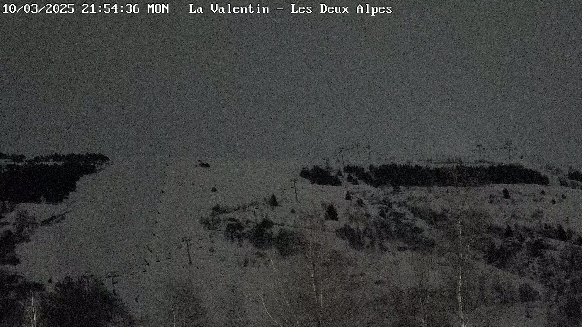 A snow-covered ski slope with sparsely wooded areas and ski lifts under a dark, overcast sky.