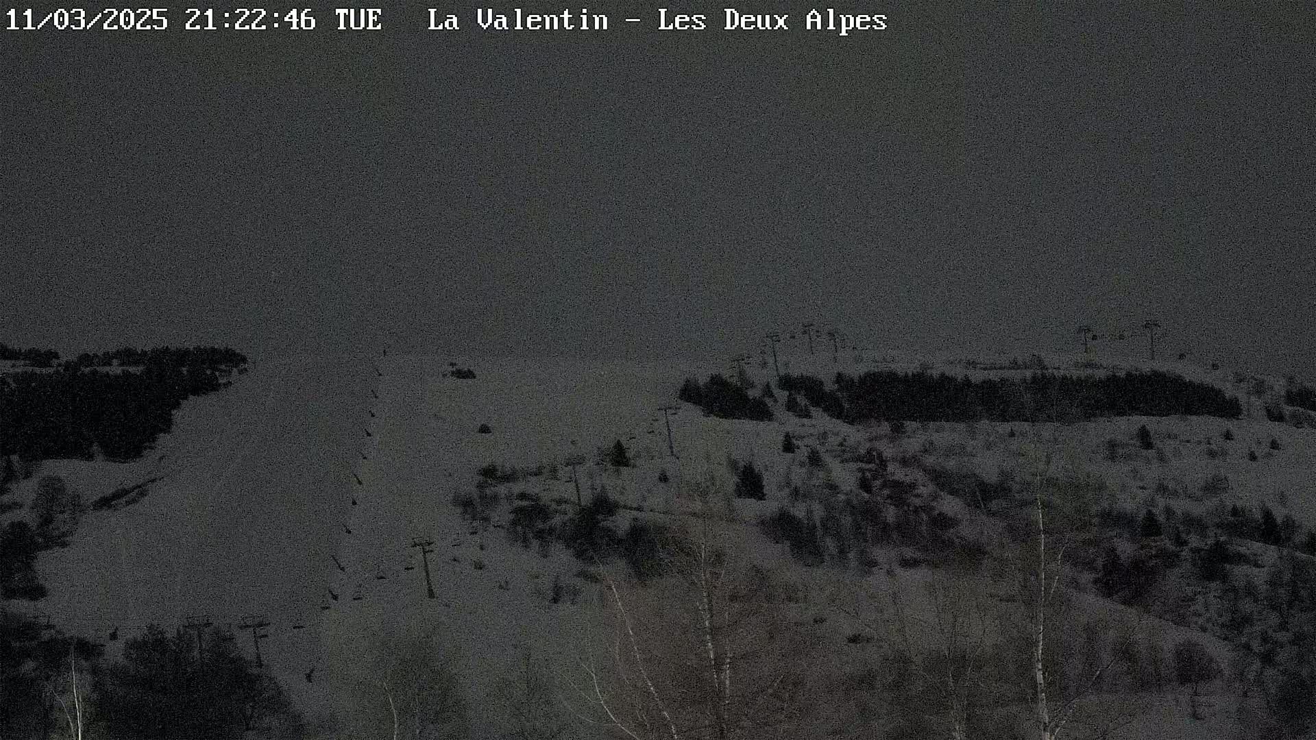 A dark, snowy ski slope with sparsely wooded areas and ski lifts is visible under a gray sky at night.