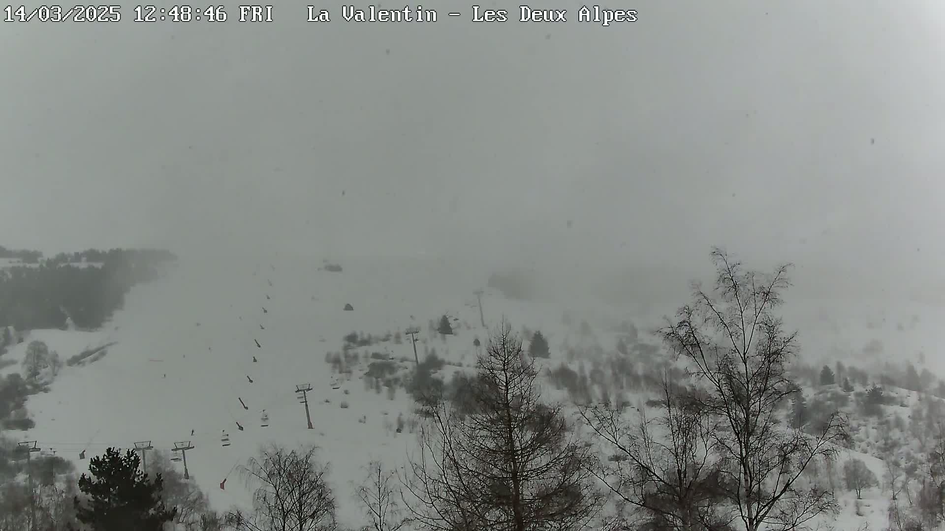 A snow-covered ski slope with sparsely wooded areas and ski lifts is visible through a heavy snowfall.