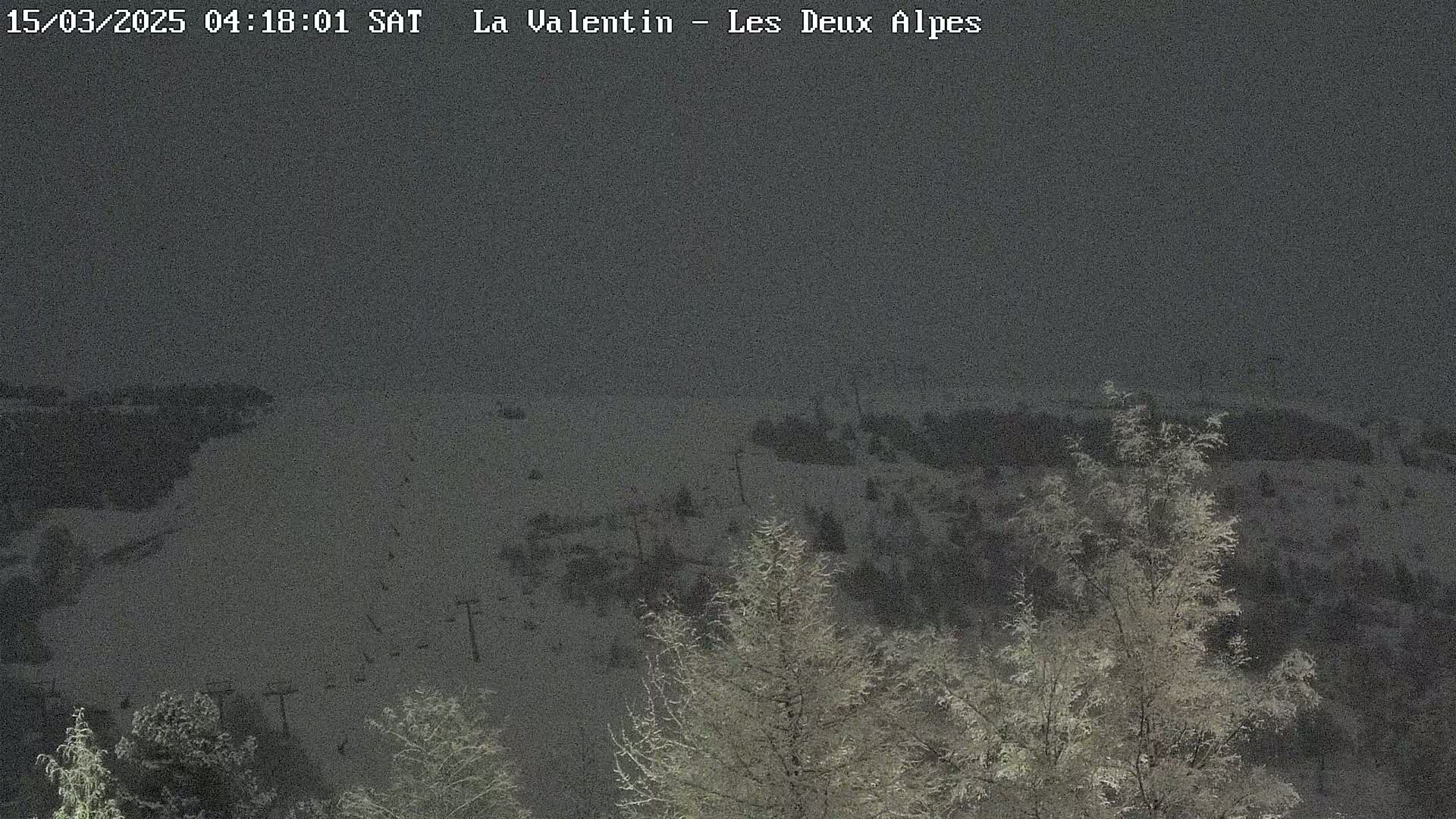 A snow-covered landscape at night, featuring frost-covered trees in the foreground and a dimly lit ski slope in the background under a dark sky.