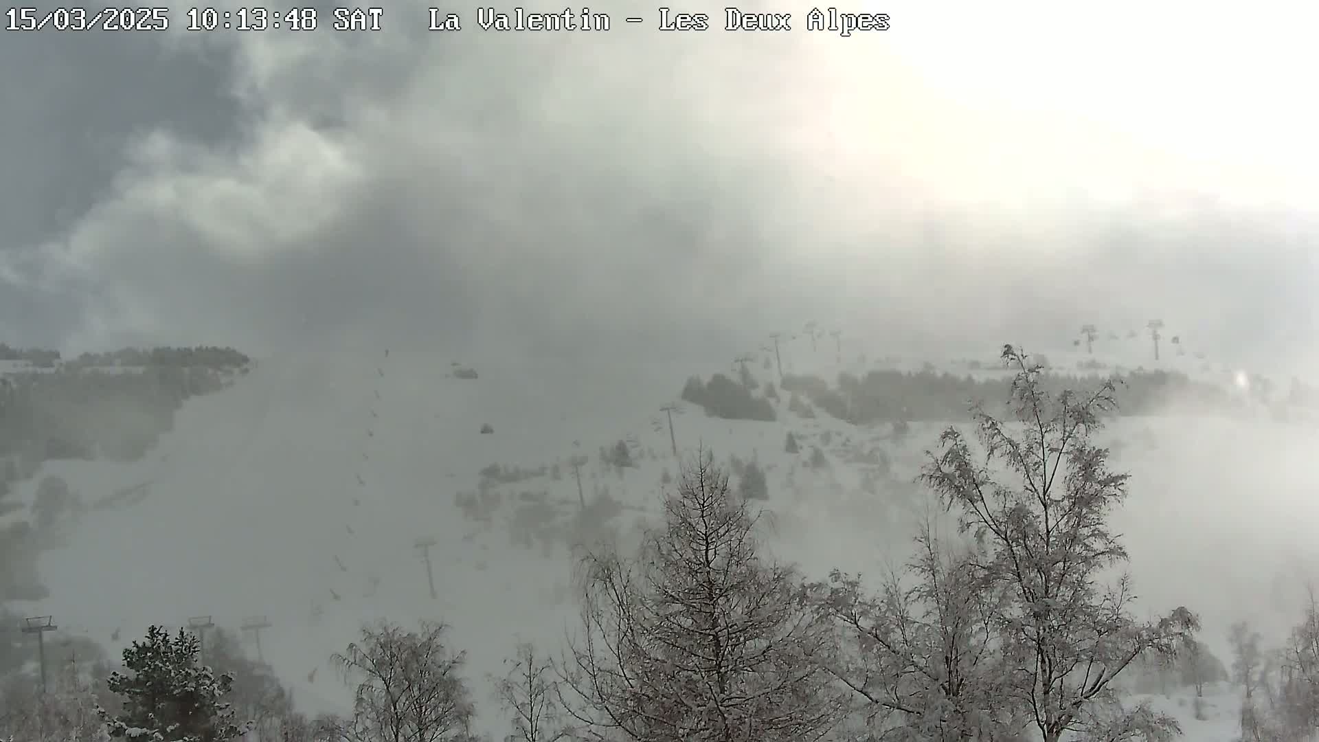 A snow-covered ski slope is mostly obscured by heavy fog or blowing snow, with snow-laden trees visible in the foreground.