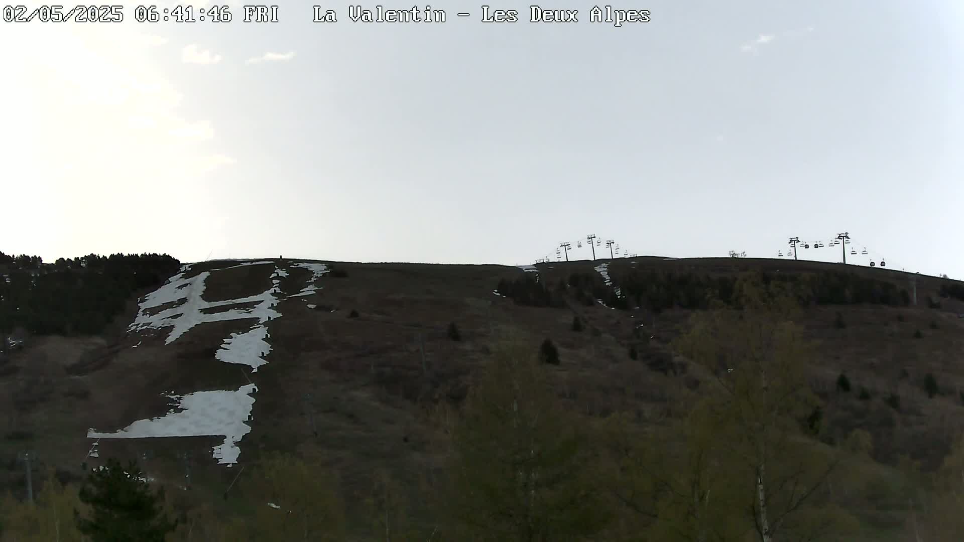 A mostly barren, brown hillside with patches of snow and several chairlifts at the top under a mostly clear, light grey sky.