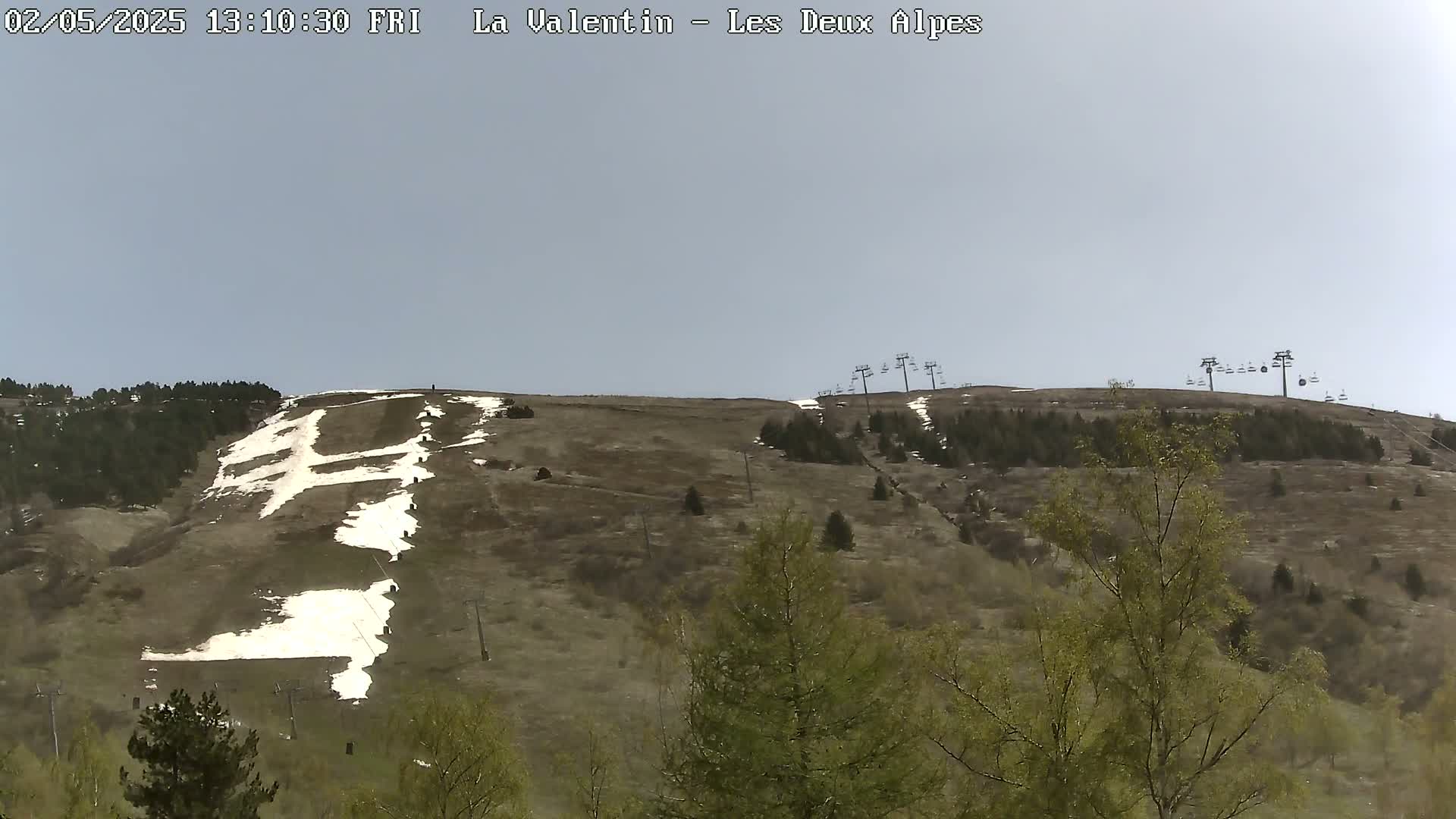 A mostly barren hillside with patches of snow, sparse trees, and chairlifts under a light gray sky.