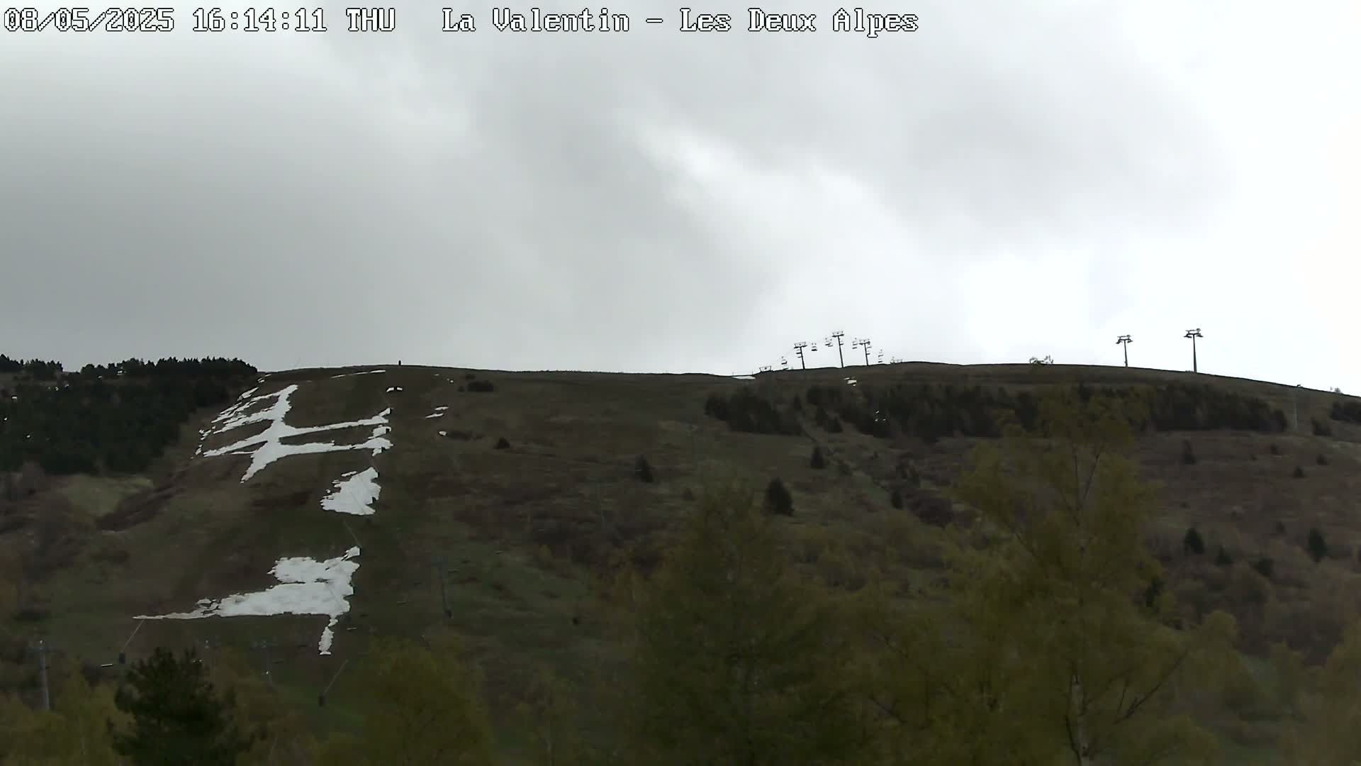 A mostly barren, green hillside with patches of snow and ski lift infrastructure is visible under an overcast sky.