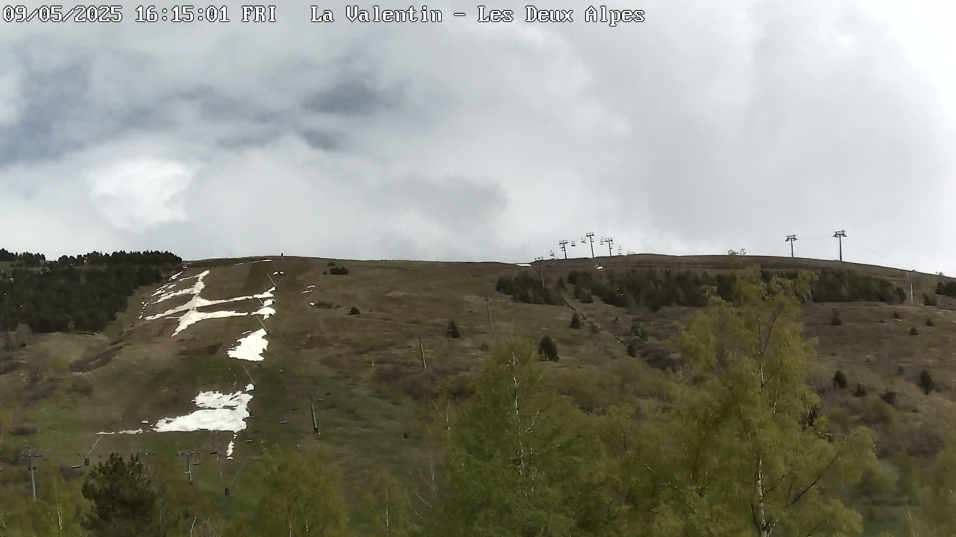 A mostly cloudy sky overlooks a hillside dotted with patchy snow, sparse trees, and chairlift infrastructure.