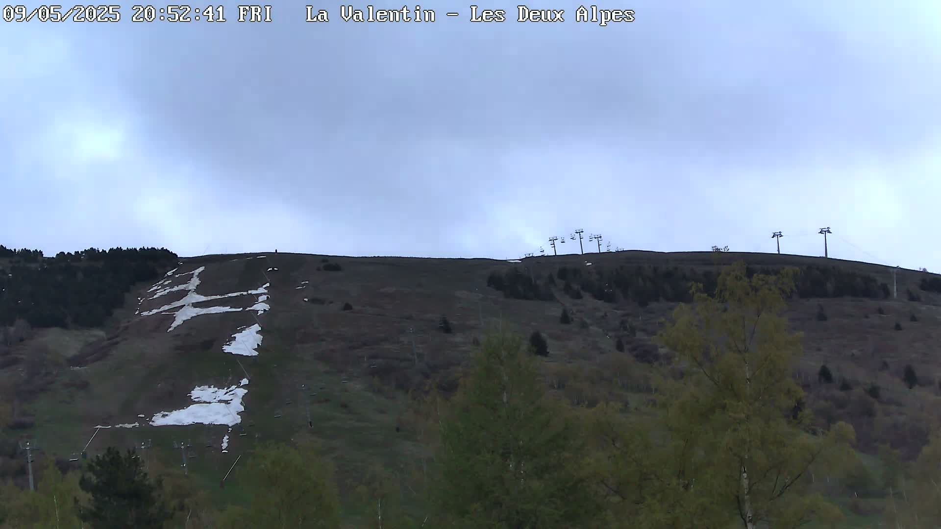 A mostly cloudy sky overlooks a hill covered in patchy snow and budding green vegetation, with ski lifts visible at the top.