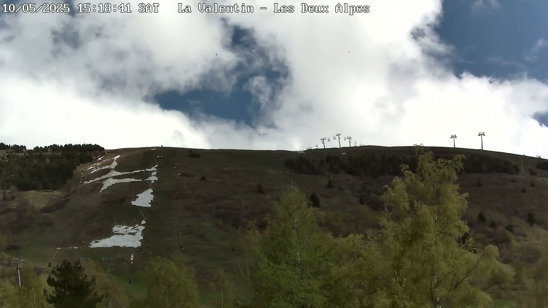 A partly cloudy sky overlooks a hillside covered in patchy snow and budding green vegetation, with ski lifts visible at the top.