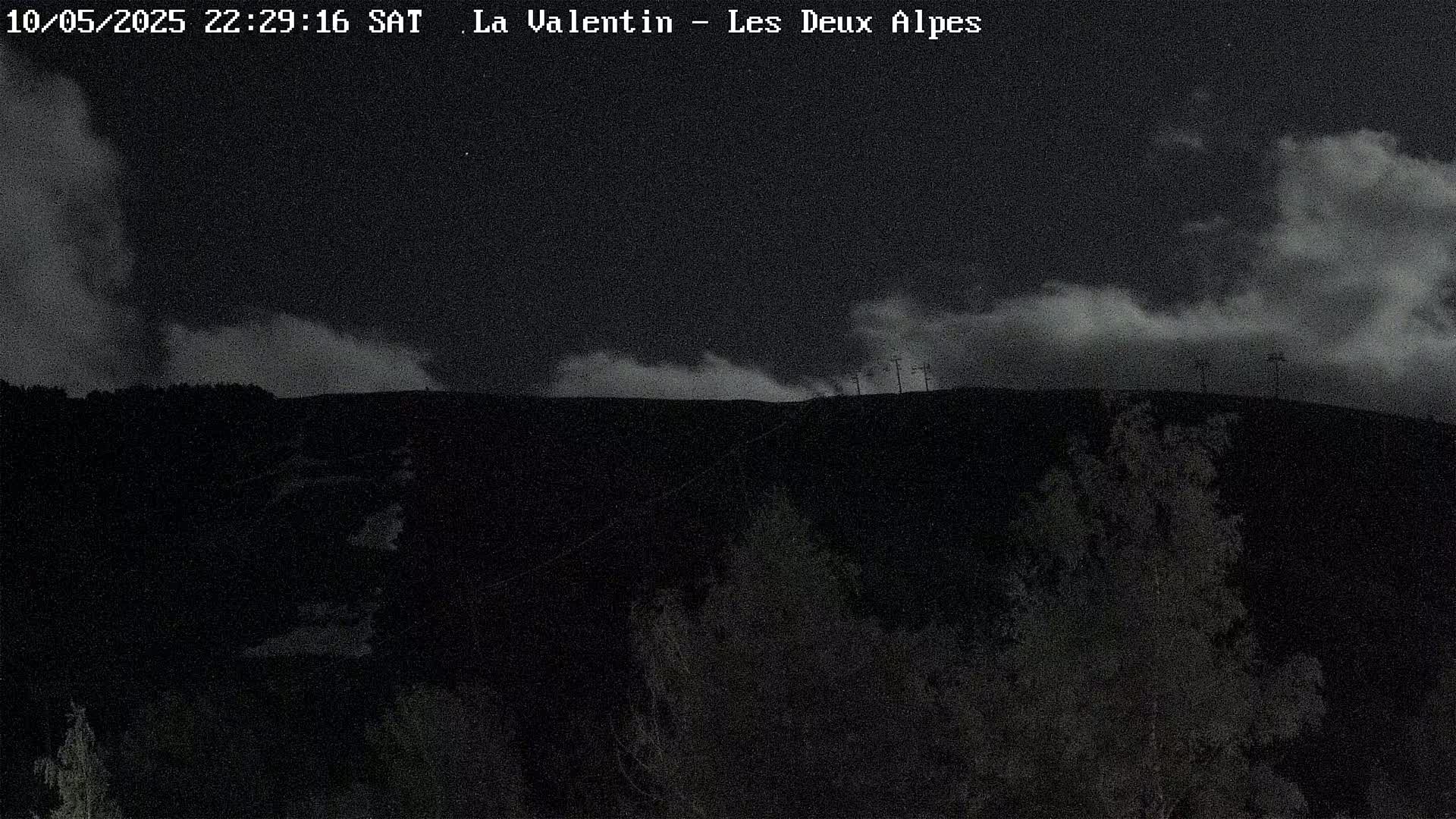 A nighttime, cloudy view of a dark hillside with sparse vegetation and a few tall structures silhouetted against the sky.