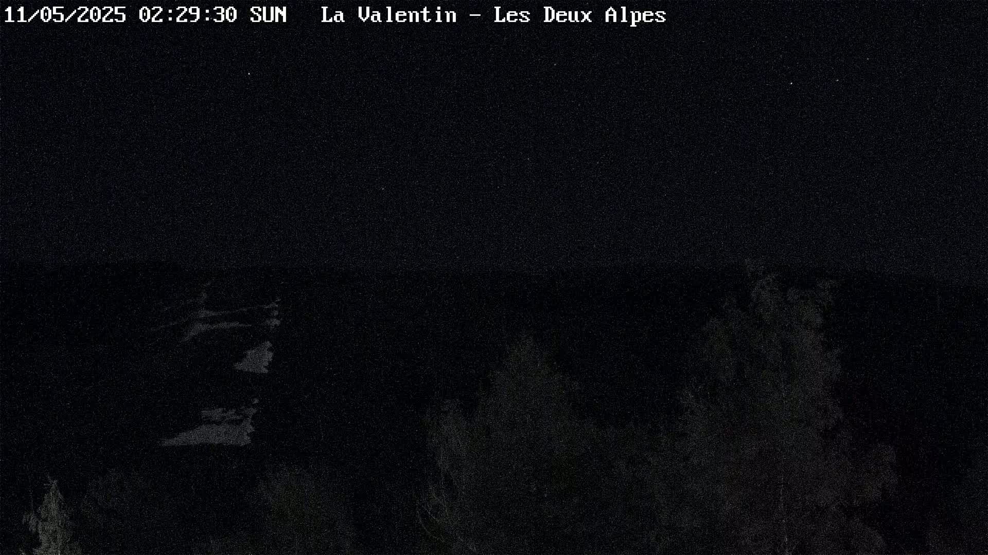 A nighttime, low-light image shows a dark, indistinct landscape possibly featuring trees under a clear, starlit sky.
