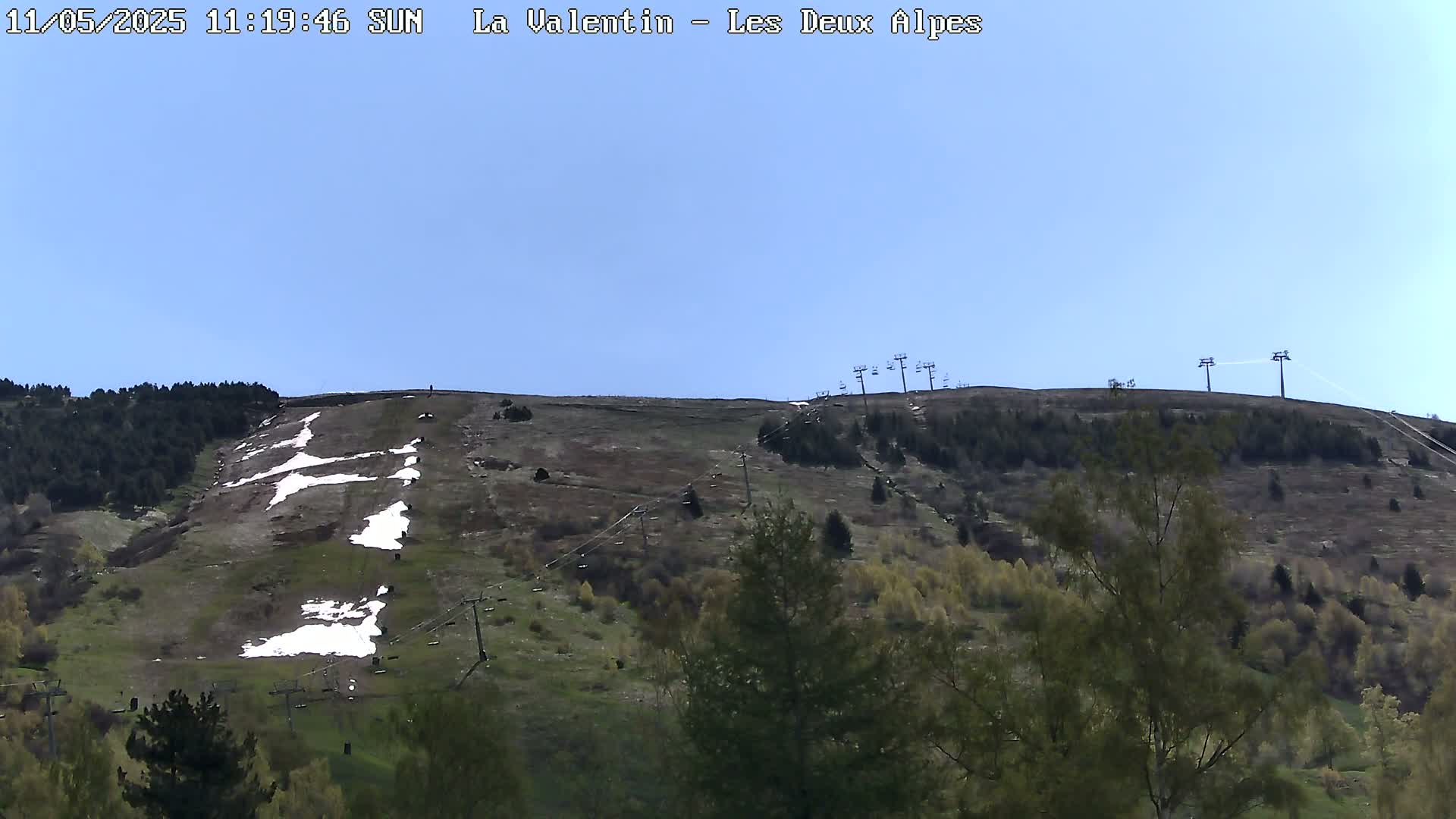 A sunny day shows a sparsely vegetated hillside with patches of snow, ski lift infrastructure, and evergreen trees in the foreground.