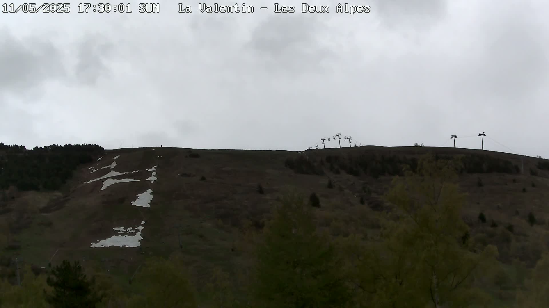 A mostly barren hillside with patches of snow, sparsely covered with dark green vegetation and ski lifts at the top, under an overcast sky.