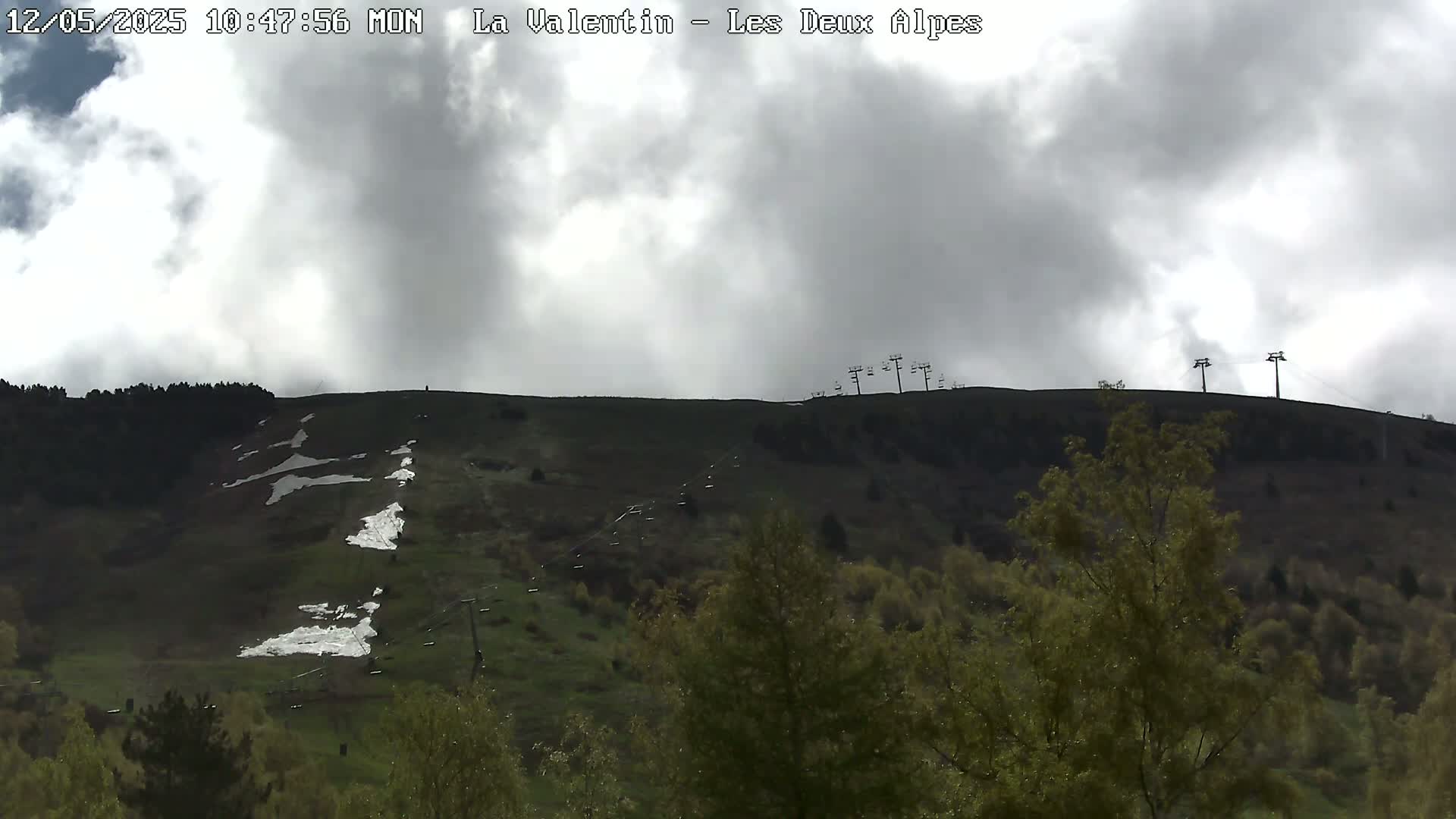 A mostly cloudy sky overlooks a grassy hillside with patches of remaining snow and ski lift towers.
