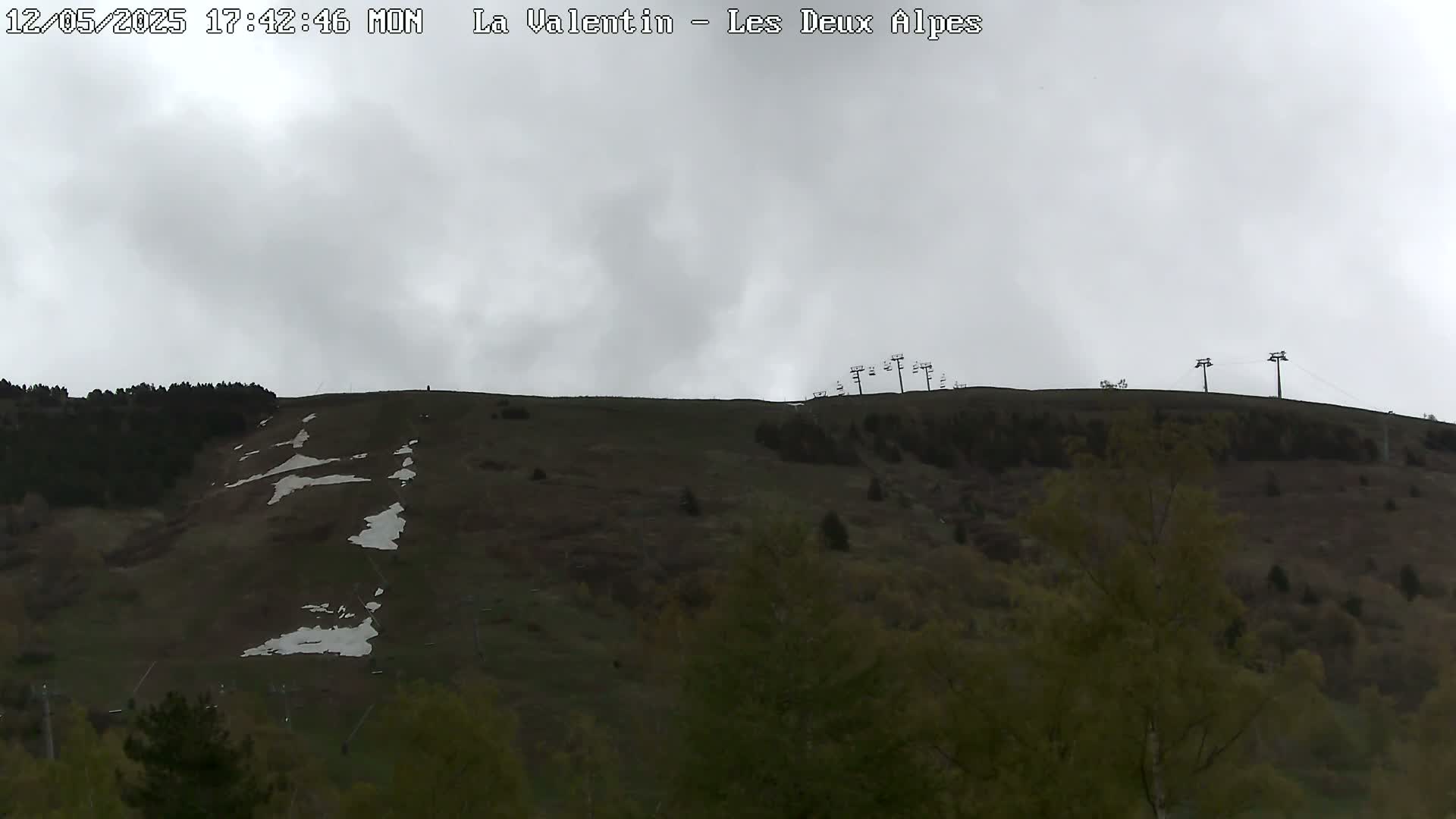 A mostly cloudy day reveals a grassy hillside with patches of remaining snow, a few trees, and ski lift infrastructure.