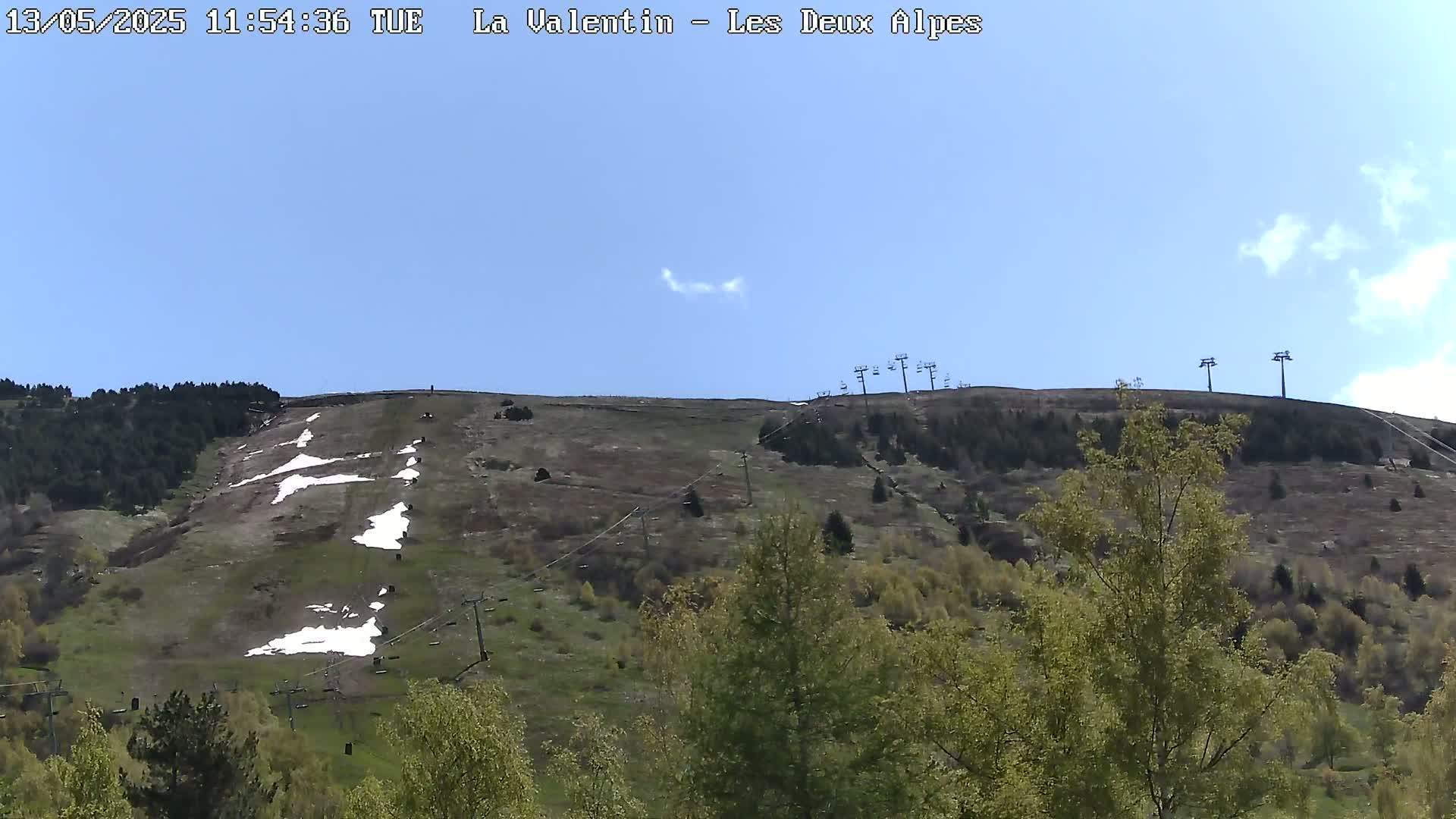 A mostly clear blue sky overlooks a grassy hillside with patches of snow and several chairlifts.