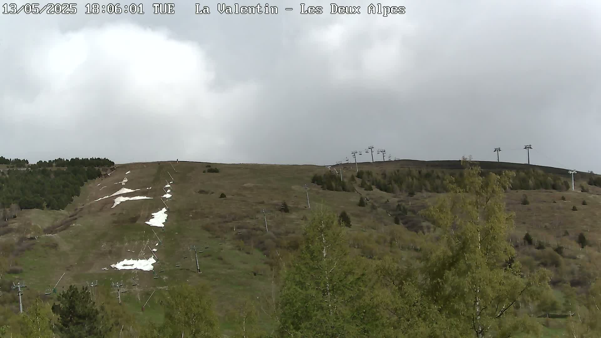 A mostly cloudy day reveals a grassy hillside with remnants of snow, ski lifts, and sparse trees.