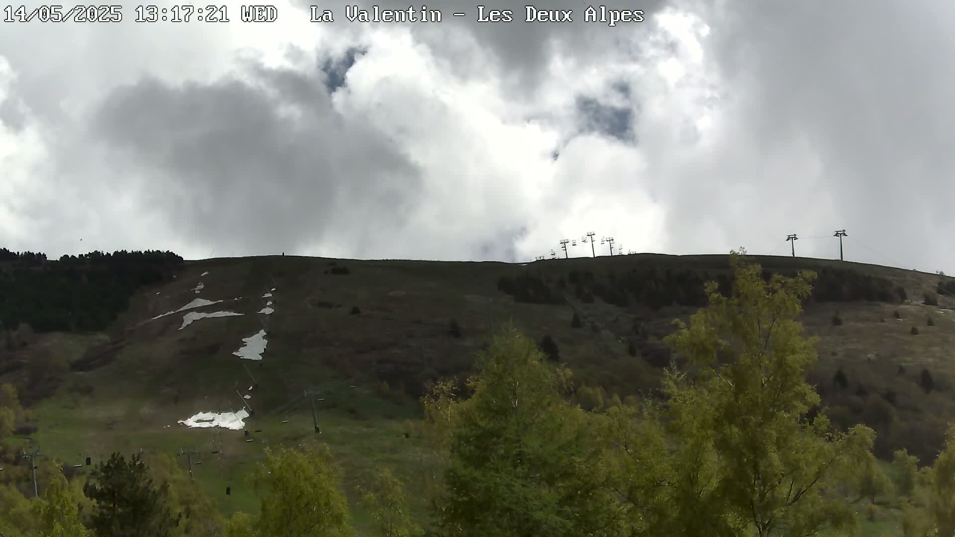 A mostly cloudy sky overlooks a grassy hillside with patches of snow, ski lifts, and green trees.