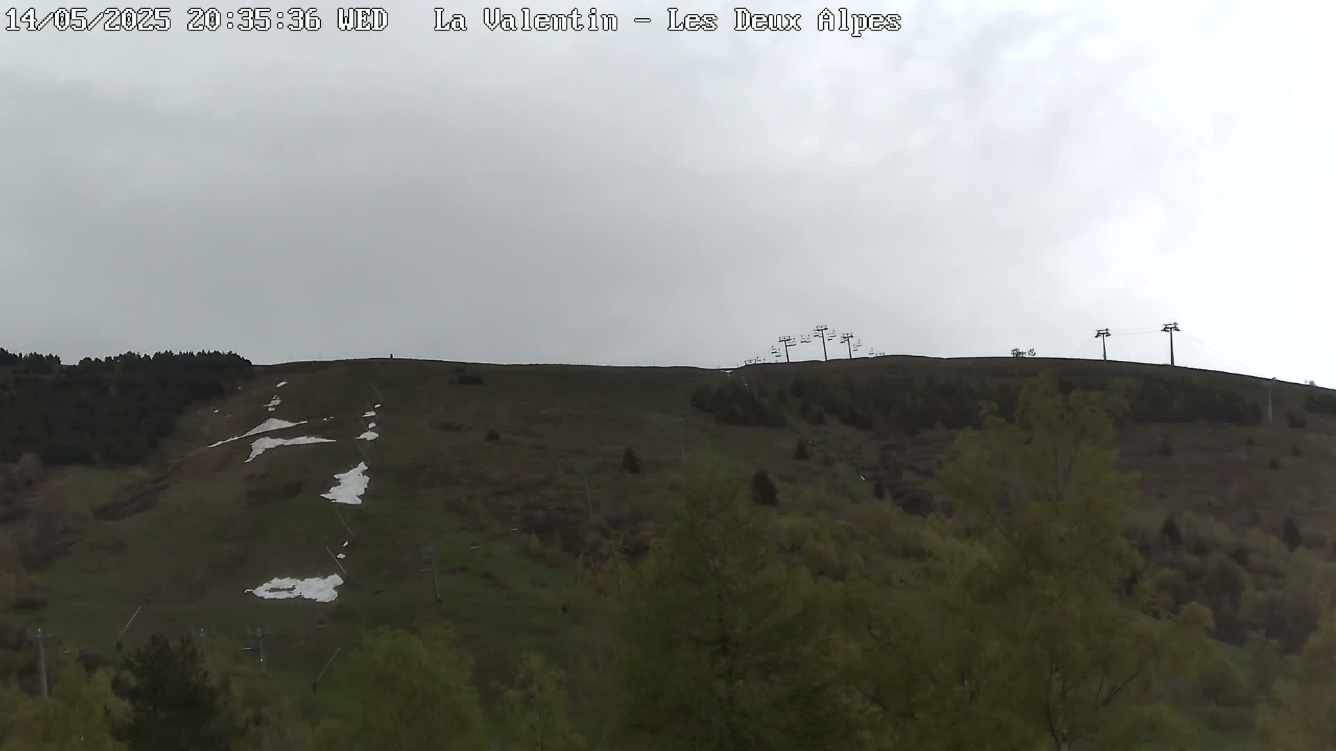 A hillside covered in low-lying vegetation and patches of snow under an overcast sky shows several chairlifts at the top.
