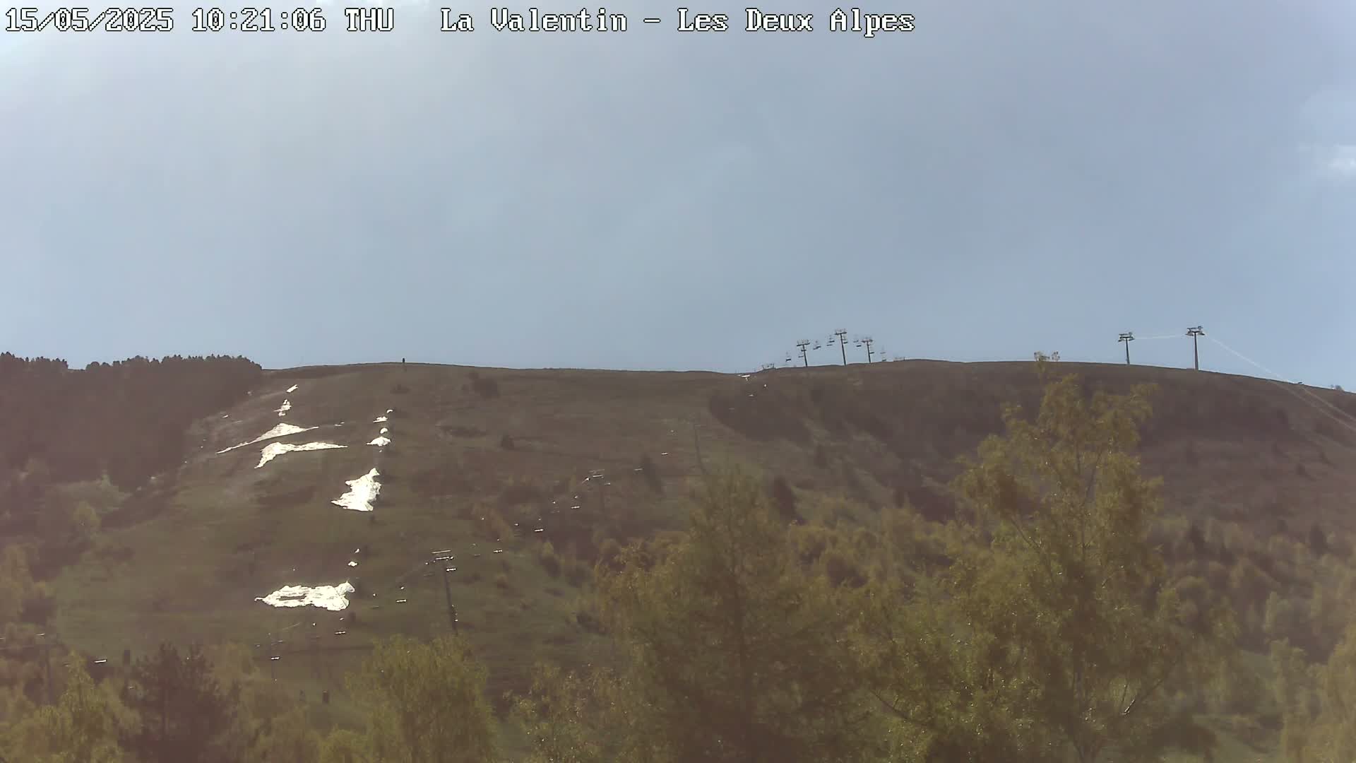 A mostly clear sky overlooks a grassy hillside with patches of snow, sparsely vegetated areas, and several chairlifts.
