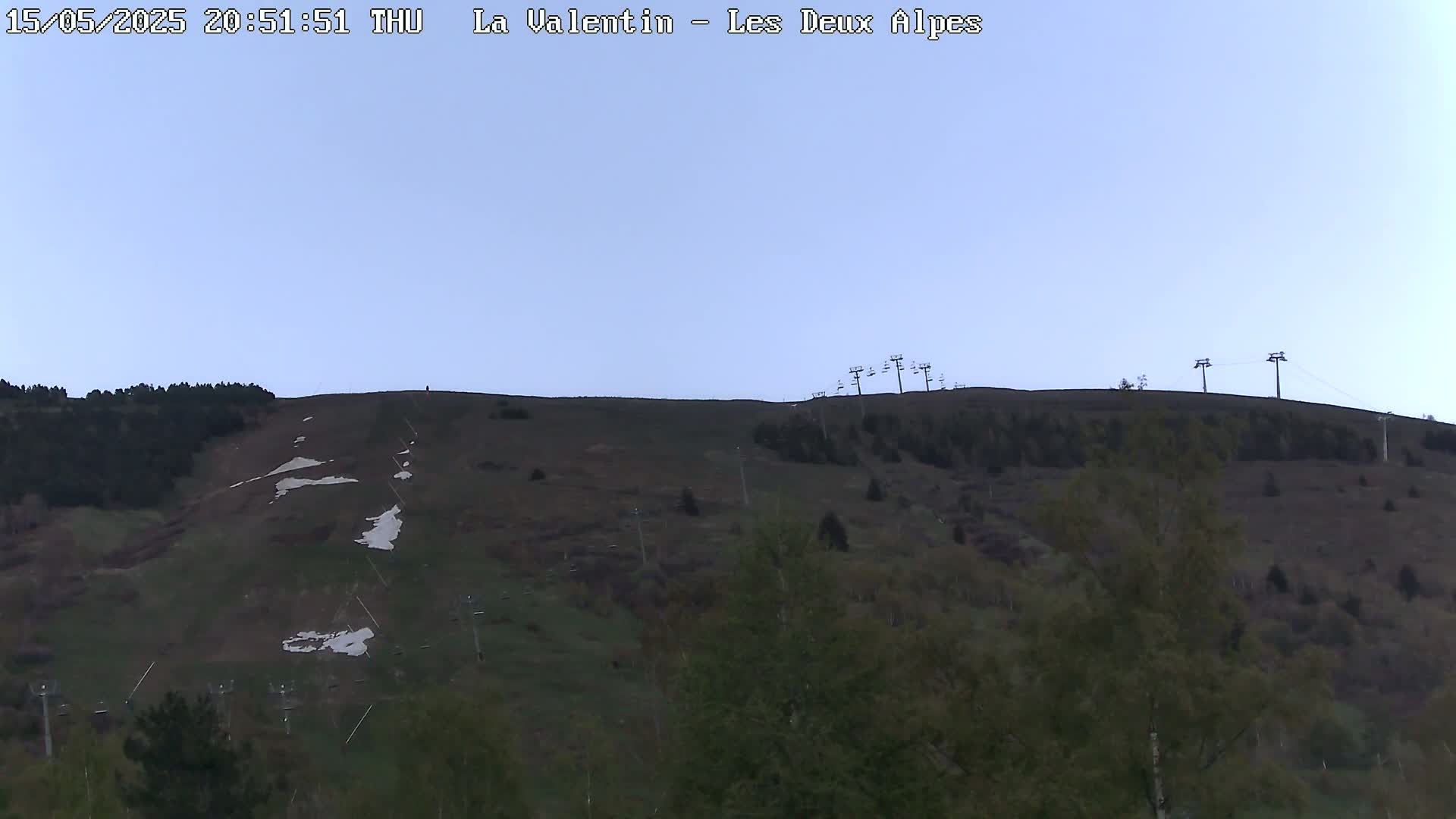 A mostly clear twilight sky overlooks a grassy hillside with sparse patches of snow and several chairlifts.