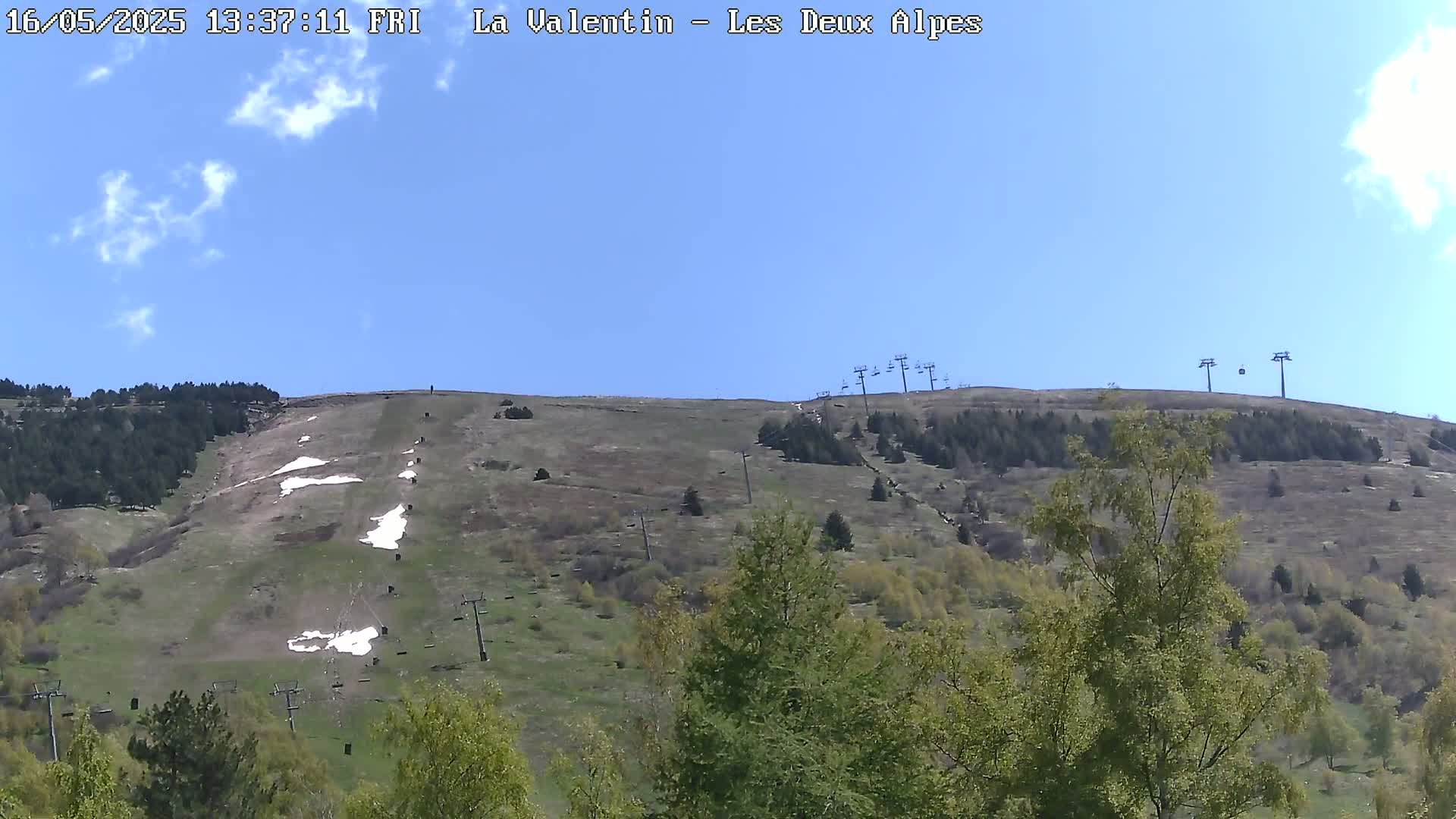 A mostly sunny sky overlooks a grassy hillside with remnants of snow, sparsely scattered trees, and several chairlifts.