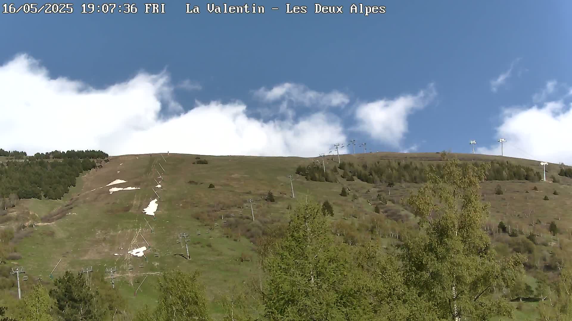 A grassy hillside with sparsely scattered trees, remnants of snow, and ski lift infrastructure under a mostly sunny sky with scattered clouds.