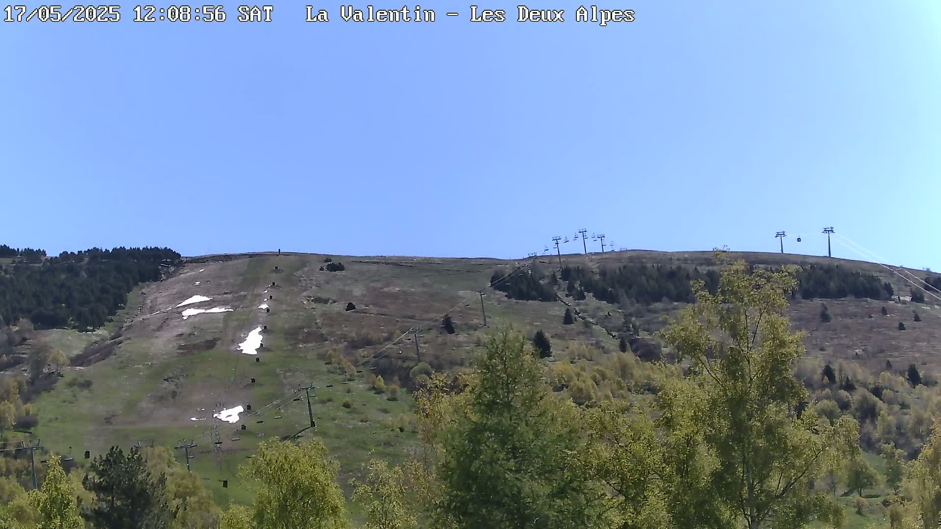 A mostly clear, sunny day reveals a grassy hillside with patches of snow, ski lifts, and sparse trees.