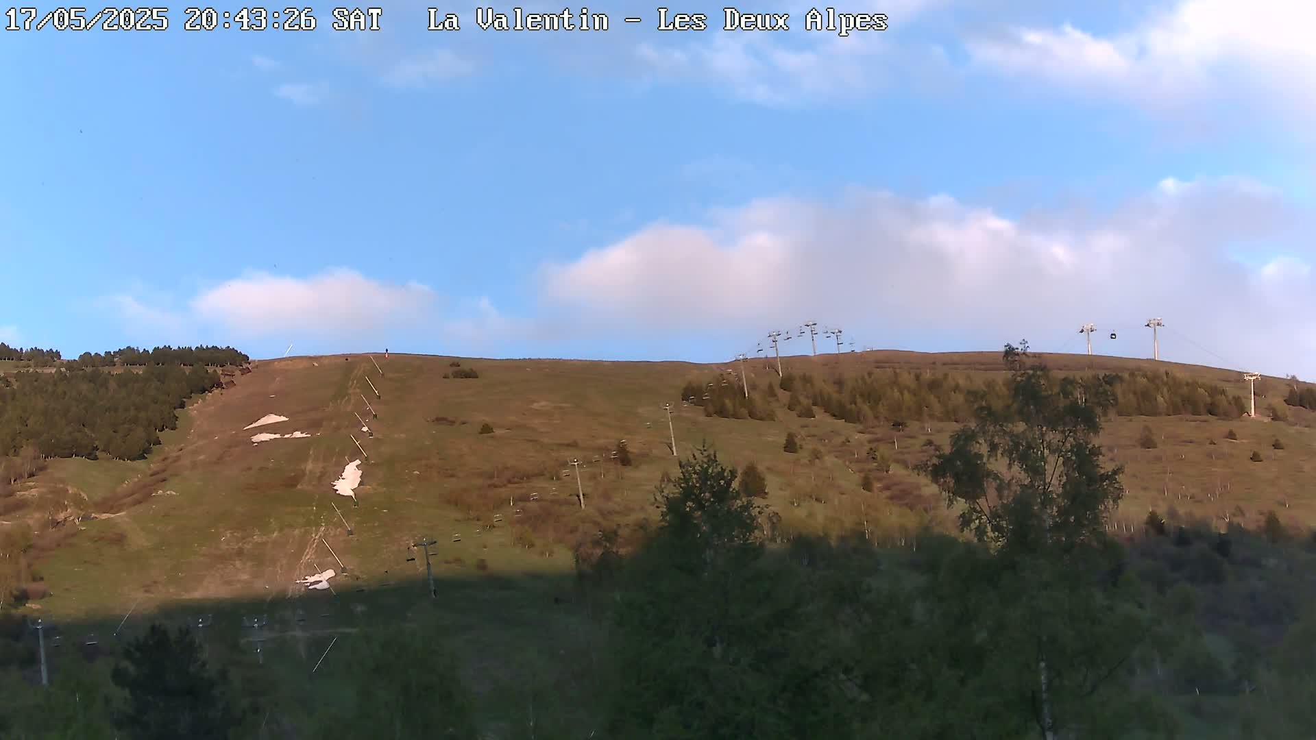 A mostly clear, blue sky with some clouds overlooks a grassy hillside with sparse trees, ski lifts, and remnants of snow.