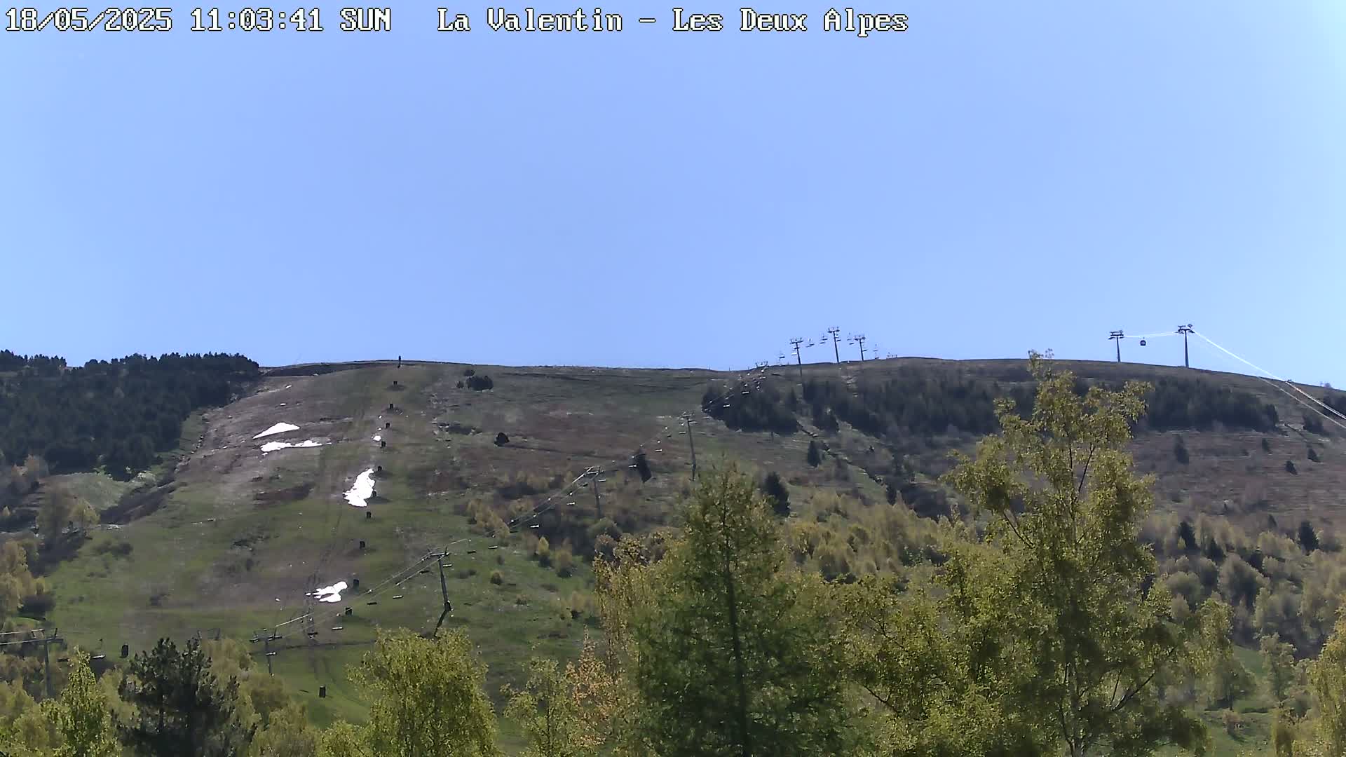 A mostly clear, sunny day reveals a grassy hillside with a ski lift and scattered patches of snow.