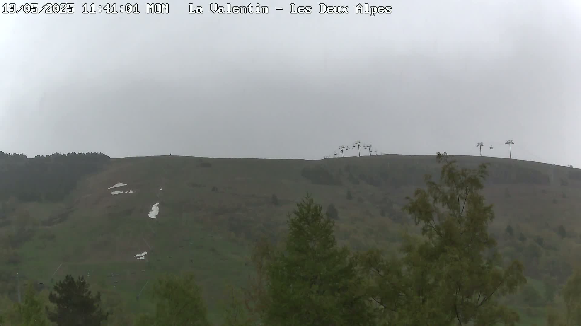 A mostly overcast view shows a grassy hillside with remnants of snow and chairlifts at the top.