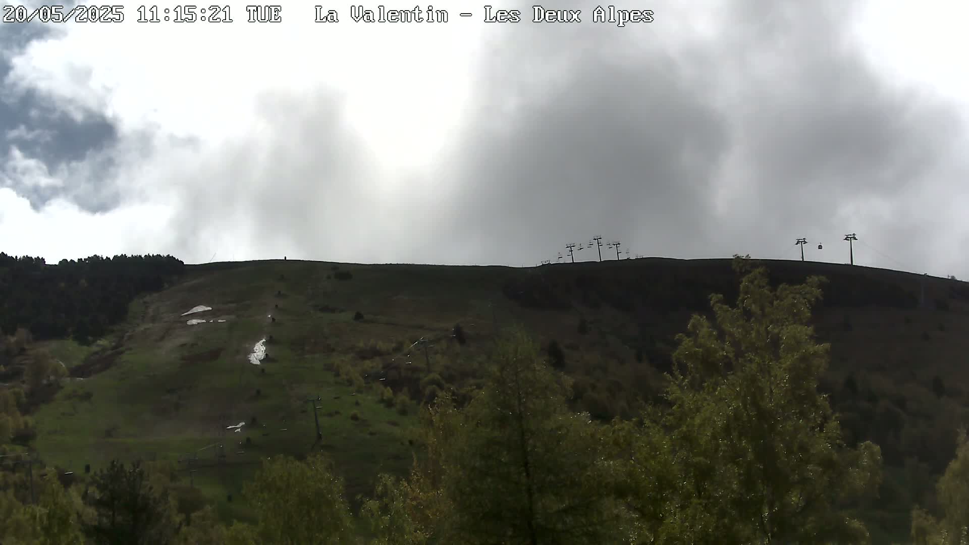 A mostly cloudy sky hangs over a grassy hillside with several chairlifts and sparse trees.