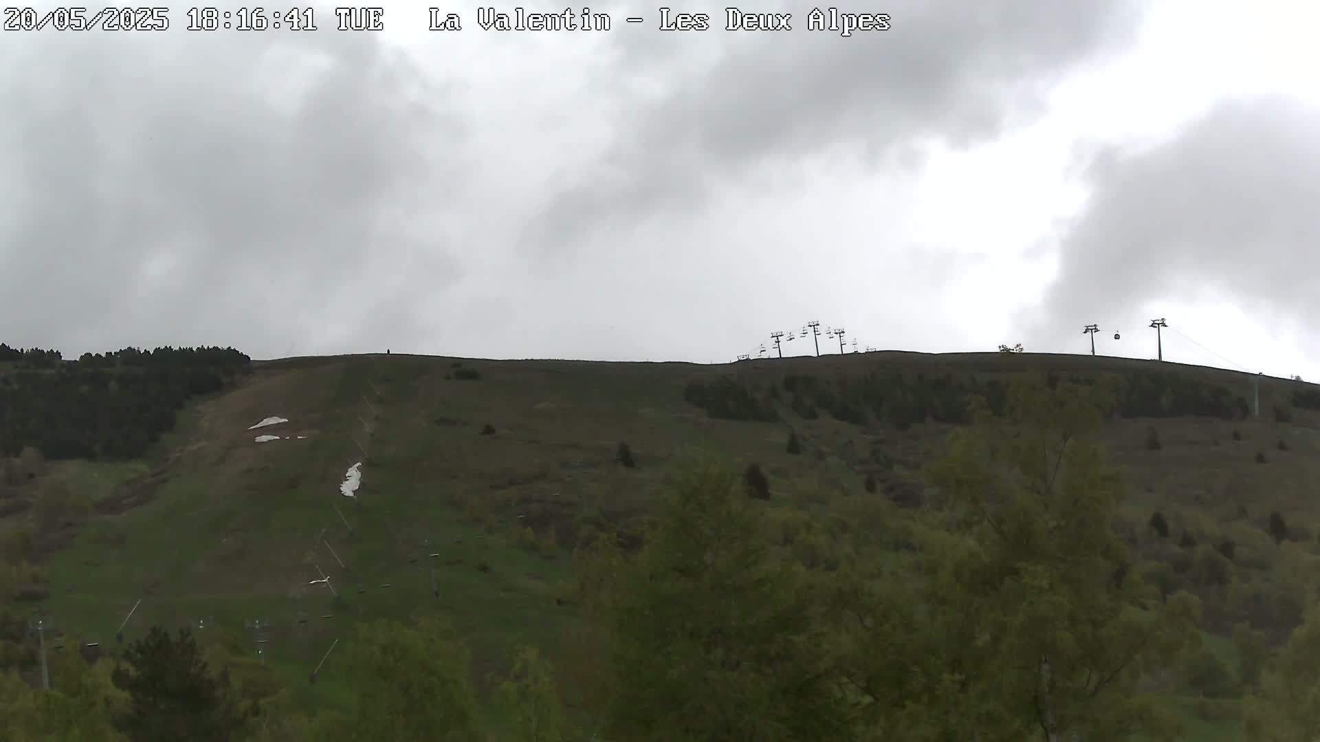 A mostly cloudy sky hangs over a grassy hillside dotted with trees and a few chairlifts.
