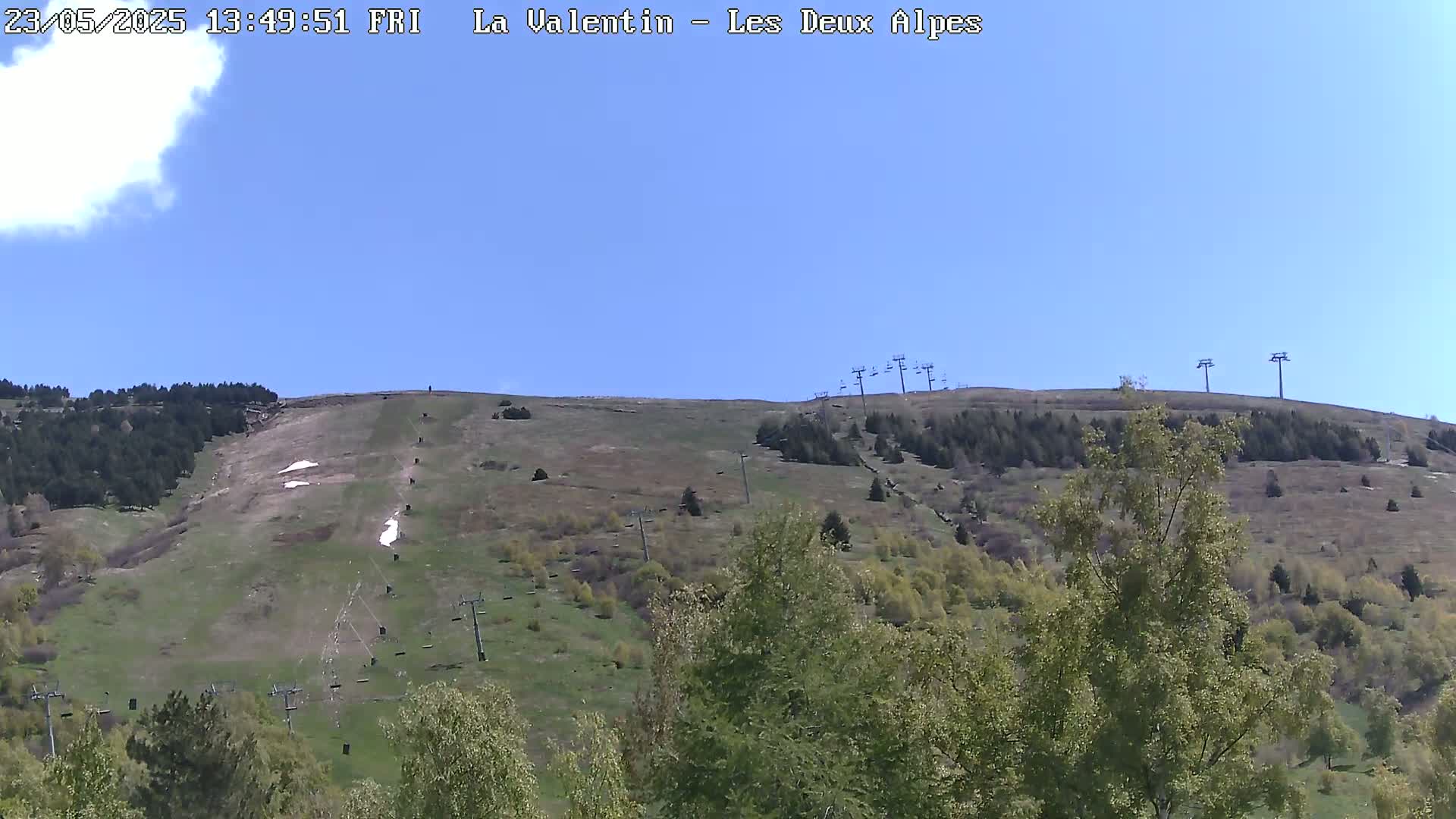 A mostly sunny day reveals a grassy hillside with sparse trees, ski lifts, and small patches of snow under a clear blue sky.