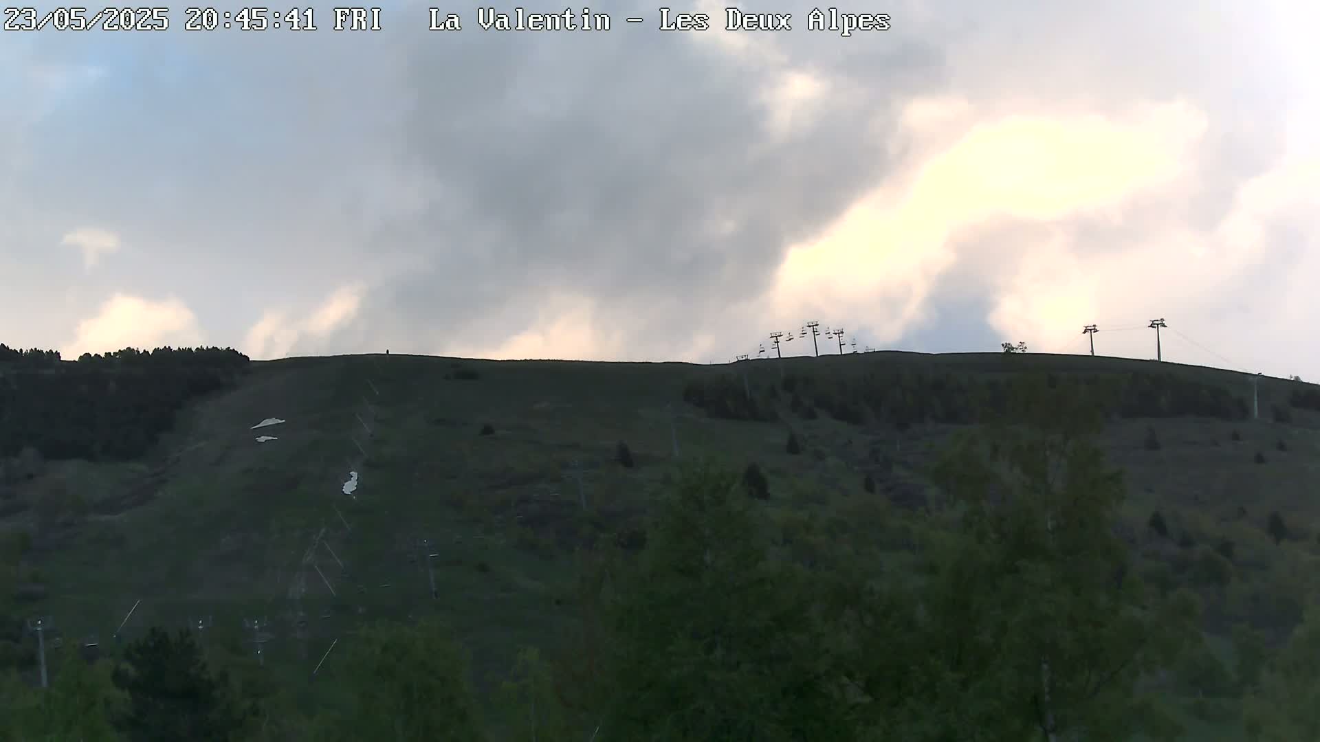 A mostly cloudy evening sky overlooks a grassy hillside with remnants of snow and chairlifts.
